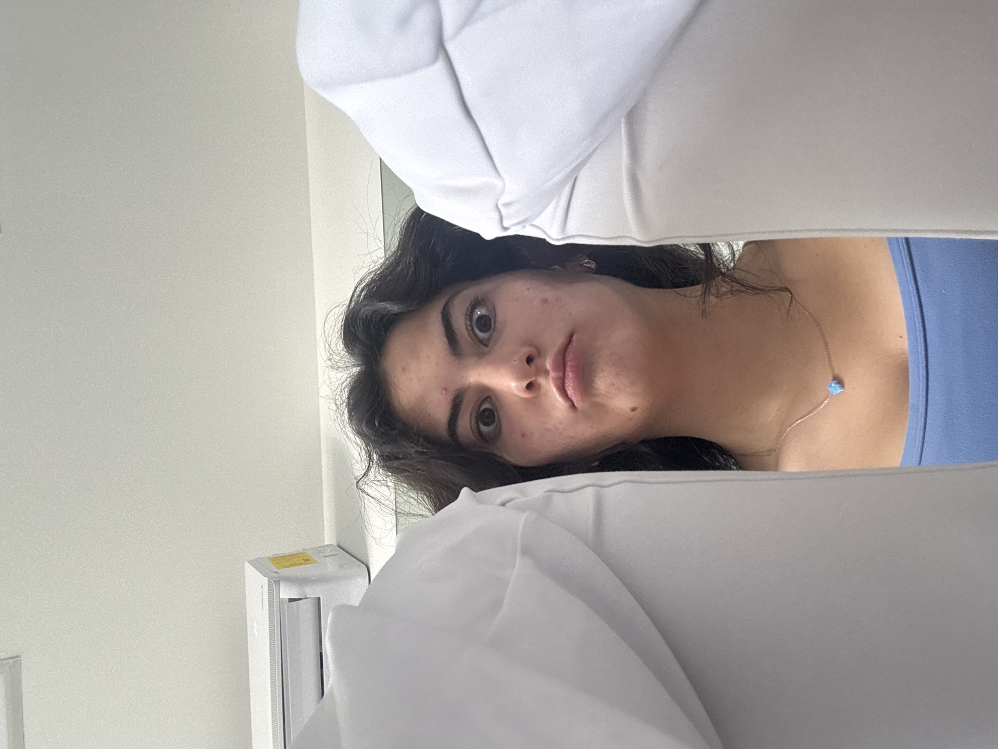 A woman is visible between two white objects, likely pillows. She has dark, curly hair and a light complexion with some visible blemishes. She is wearing a blue top and a silver necklace with a light blue pendant. The background includes a white wall and what appears to be an air conditioning unit. There is no information within the image to identify the location as San Andrés Cholula, Mexico.