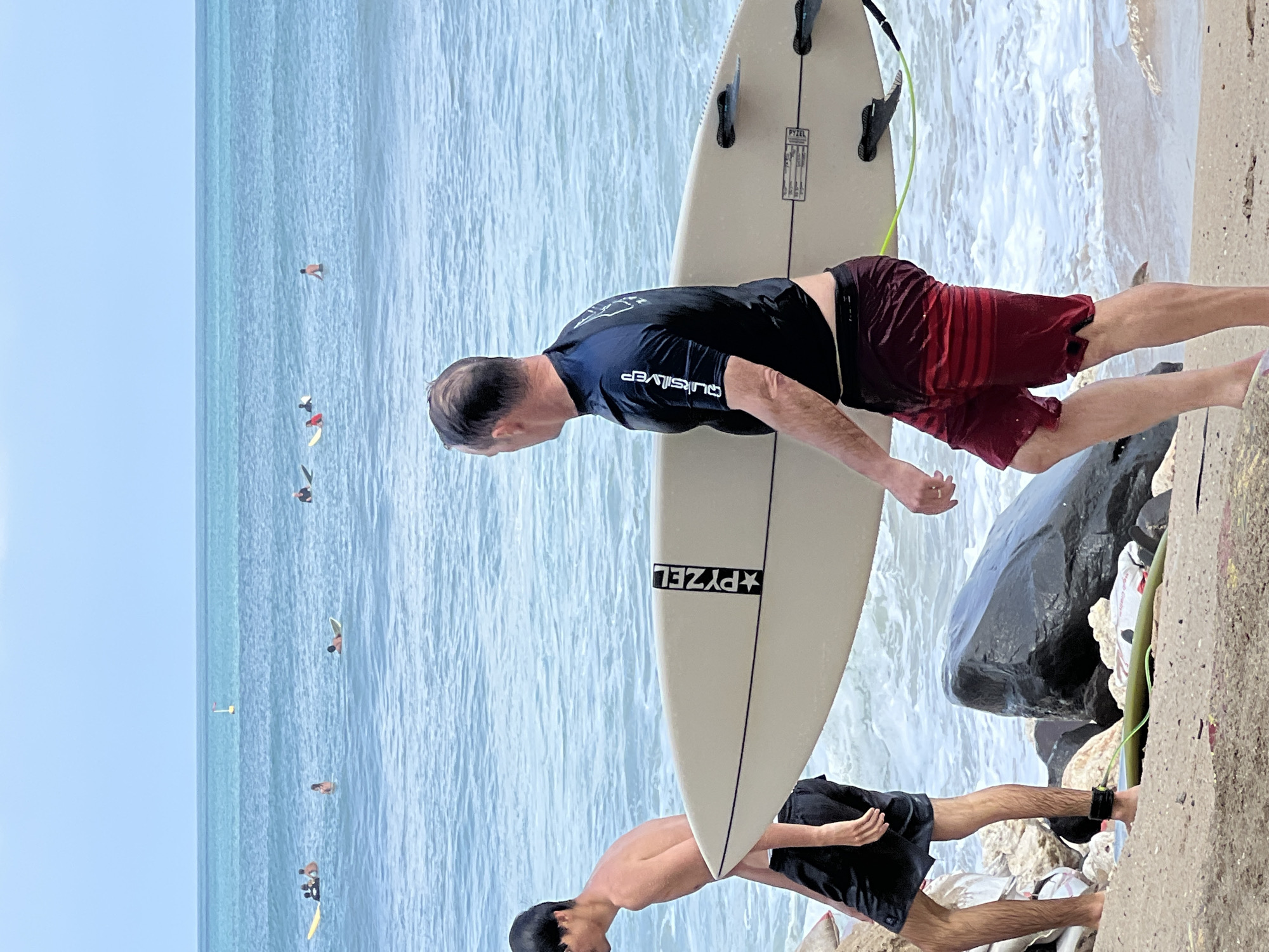 Two men are walking on the beach in Kuta, Indonesia, carrying a surfboard. The man in the foreground on the right is wearing a black rash guard and maroon board shorts, and carrying the surfboard. The man on the left is wearing black board shorts and is shirtless. The ocean is calm with a few surfers in the distance. The sky is overcast.