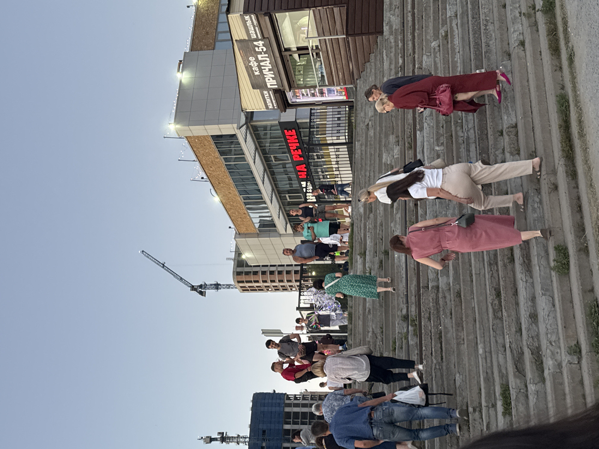The image captures a scene of people walking and standing on concrete stairs outdoors, likely in a public area. The people are diverse in age and attire, suggesting a casual gathering or a stroll. Some individuals are seen in conversation or walking together, while others appear to be moving independently.
In the background, a modern building complex is visible, featuring glass facades and architectural details. A prominent sign in red neon letters reads "НА РЕЧКЕ," which translates to "ON THE RIVER." Another sign above a shop says "КАФЕ ПРИЧАЛ-54" and "ШАШЛЫК," indicating a cafe named "Prial-54" that serves shashlik (kebabs). The presence of these signs, along with the outdoor setting and what appears to be evening light, suggests a vibrant urban environment, possibly a waterfront area in Novosibirsk, Russia.
Construction cranes are visible in the distance, hinting at ongoing development or urban renewal in the area. The sky is clear and bright, suggesting fair weather. The overall mood appears relaxed and social, with people enjoying the outdoor space. The composition of the image, with the stairs leading upwards and the building and sky forming the backdrop, creates a sense of depth and scale. The orientation of the image is rotated, with the ground appearing on the left side and the sky on the right.