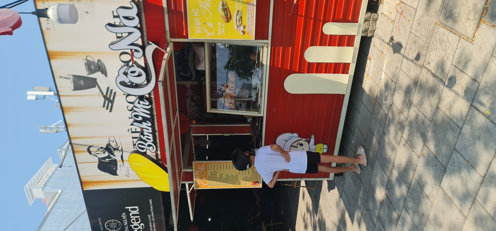 A person stands in front of a food stall, likely a Bánh Mì vendor, in the city of Đà Nẵng, Vietnam. The stall is red with white accents and features signage for "Bánh Mì Cô Na" and "Coffee." A menu board is visible, listing various Bánh Mì options and prices. The person appears to be examining the menu. The location is near 25 Hồ Nghinh, Đà Nẵng, as indicated on the stall's signage. The environment includes paved ground and possibly a building structure in the background.