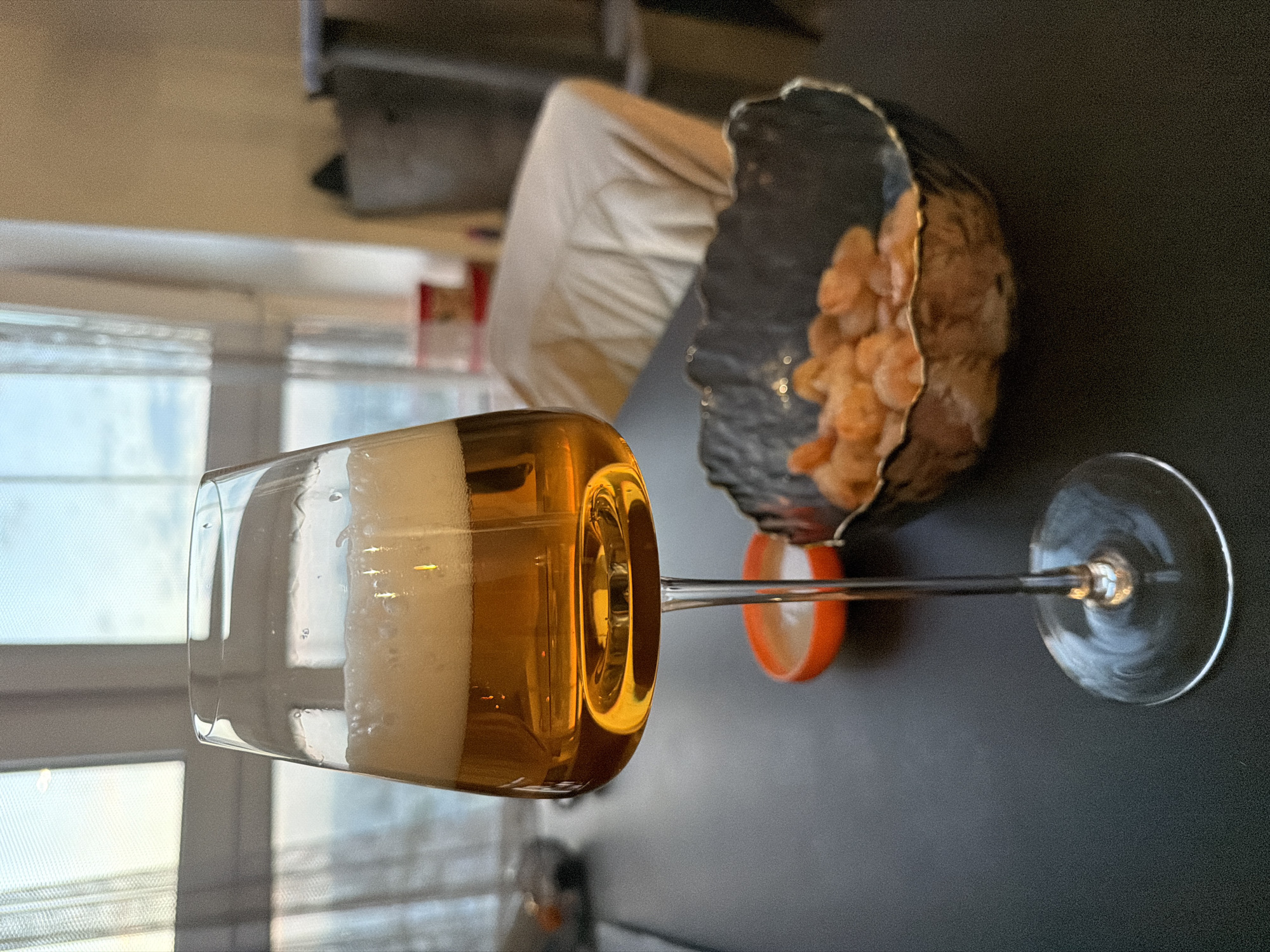 The image shows a glass of beer and a bowl of small shrimp or prawns on a dark surface. The beer glass, filled with a frothy, golden beverage, is angled to the left. Adjacent to it is a roughly hewn, dark-colored bowl containing a quantity of small, cooked shrimp. An orange object is visible under the bowl between it and the glass. In the background, there is a blurred scene of what appears to be an interior space with furniture. The location is unspecified.