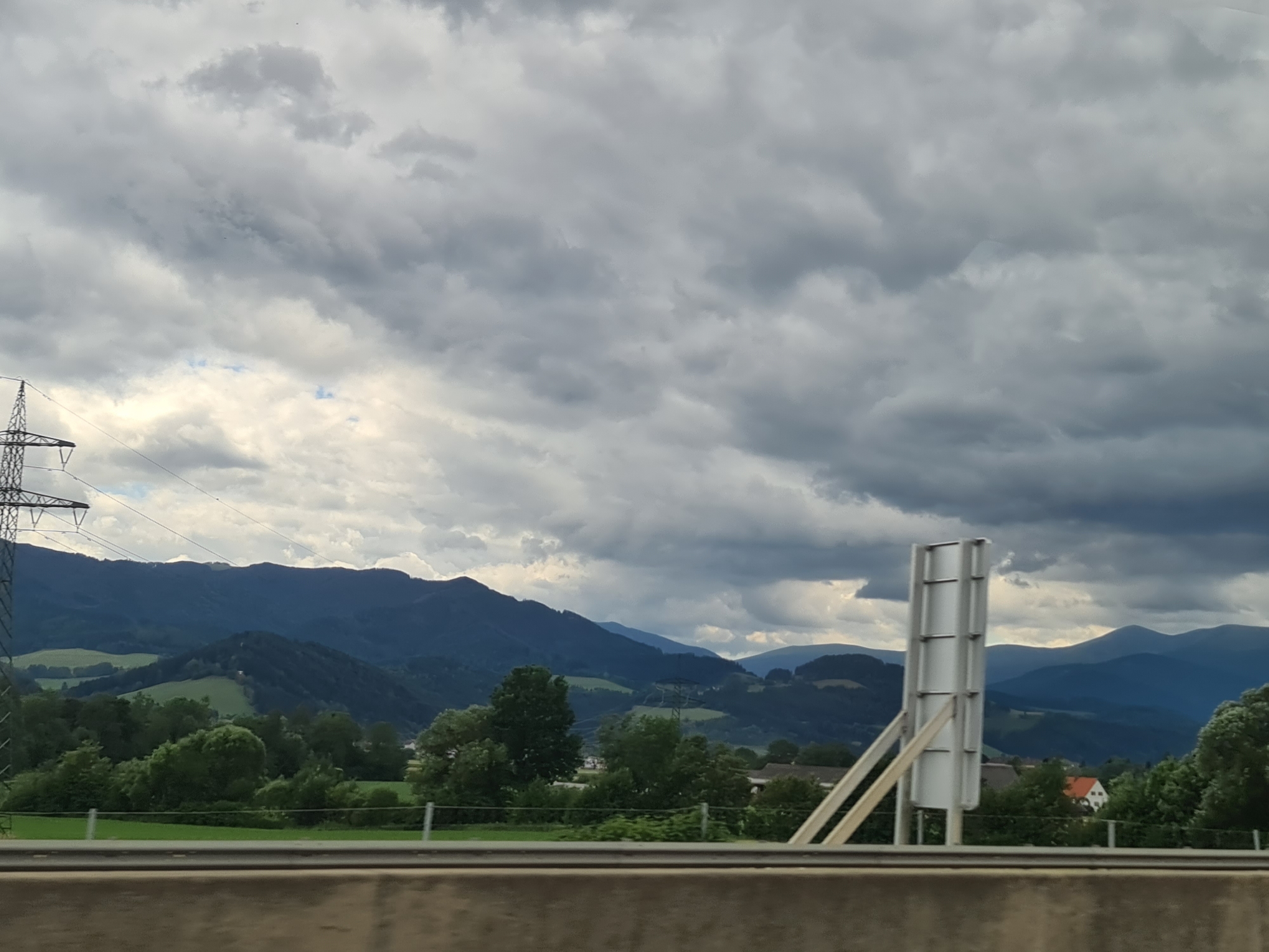 The image depicts a mountainous landscape under a cloudy sky, viewed from a highway or elevated road. In the foreground, there is a concrete barrier. To the left, a tall, lattice-like electricity pylon is visible. The middle ground features rolling green hills, dense green trees, and some scattered houses, including a prominent orange-roofed house. In the background, layers of dark blue mountains recede into the distance. A large, white, rectangular sign on a metal support structure is positioned in the right-middle of the frame, obscuring a portion of the landscape behind it. The sky is filled with dramatic, dark gray clouds, suggesting an overcast or approaching storm. The overall impression is one of a rural, mountainous region, likely in Austria given the reference to Hautzenbichl. There are no people or animals visible in the frame.