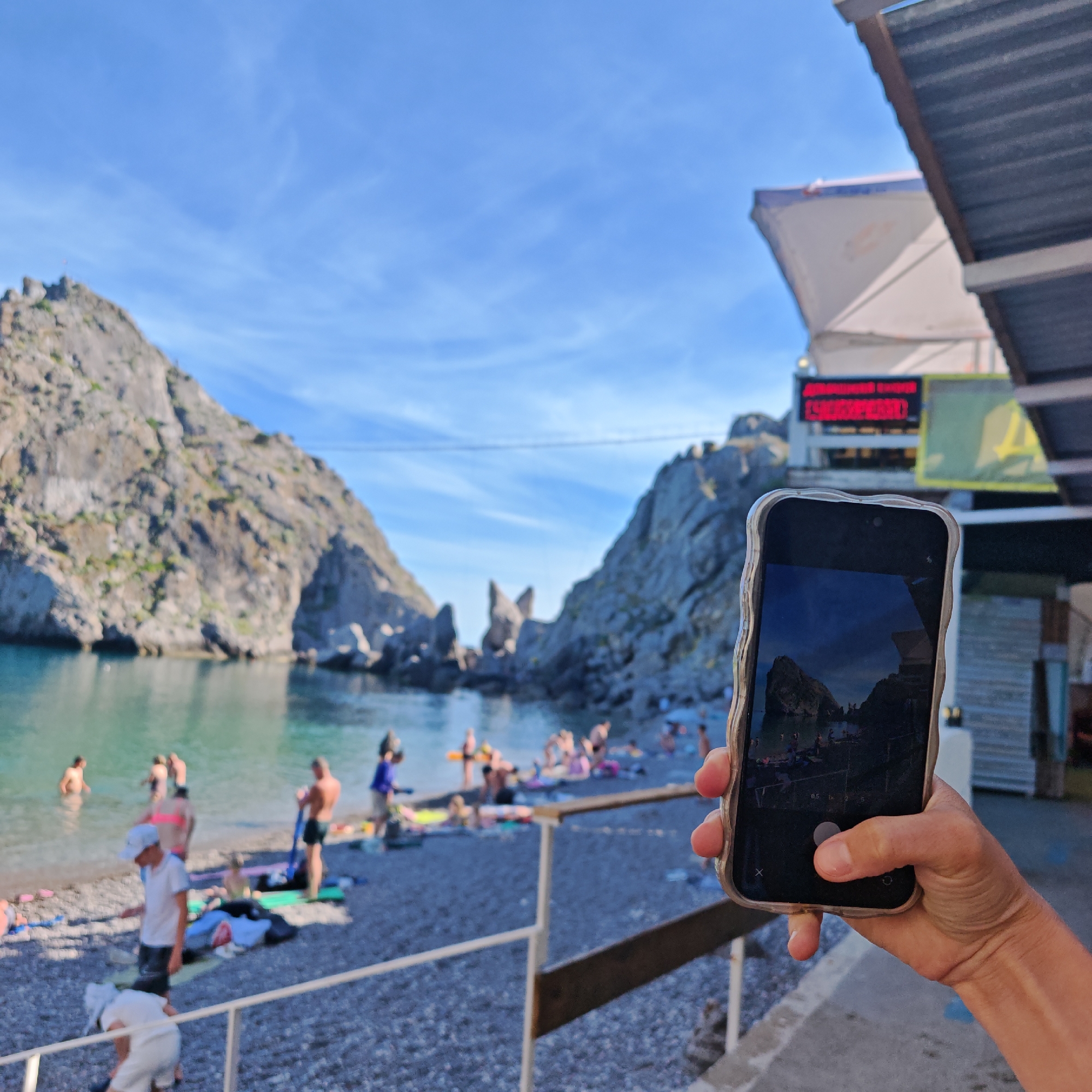 This media file depicts a sun-drenched beach scene, likely captured on a clear day. The focus is a hand holding a smartphone, which is photographing a rocky cove. In the foreground, a person's hand is visible, holding a smartphone in a textured, silver case. The phone screen displays a reflection of the same beach scene, albeit with a darker, more saturated tone, showing people on the beach and the surrounding cliffs.
The wider scene, visible both directly and on the phone screen, features a picturesque cove with imposing, rocky cliffs that drop into calm, turquoise water. A pebble beach lines the shore, where numerous people are engaged in various beach activities. Some are wading in the water, while others are relaxing on towels or mats. Children are also present, contributing to the lively atmosphere.
The time of day appears to be late afternoon or early evening, suggested by the warm sunlight casting shadows and the golden hue on some surfaces. The sky is a vibrant blue with wispy white clouds.
In the background, beyond the beach, a building with a white awning is visible, adorned with a red LED sign. The text on this sign is partially obscured but appears to be illuminated. Another sign with a large yellow character is also present.
The overall mood is one of leisure and vacation. The setting evokes a sense of natural beauty and relaxation, common to coastal resort areas. The act of taking a photograph within the photograph adds a layer of meta-commentary on the experience of capturing memories.