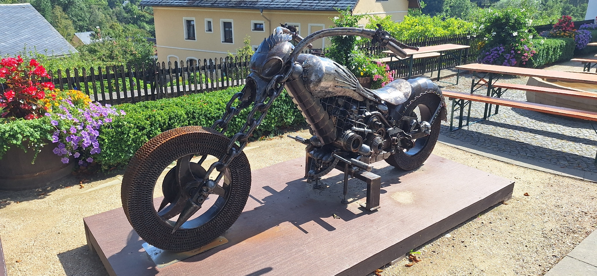 The image depicts a metal sculpture of a motorcycle displayed outdoors in Zschopau, Germany. The sculpture is constructed from various metal components, including chains, gears, and what appears to be re-purposed industrial parts. It is positioned on a raised platform. In the background, there is a fence bordering a garden with colorful flowers, and behind that, a building with a light-colored facade and windows. Several picnic tables with wooden benches are visible to the right of the motorcycle sculpture, set up on a cobbled area. The overall environment suggests a public outdoor space, possibly a beer garden or a park area within the city. There are no people visible in the image.