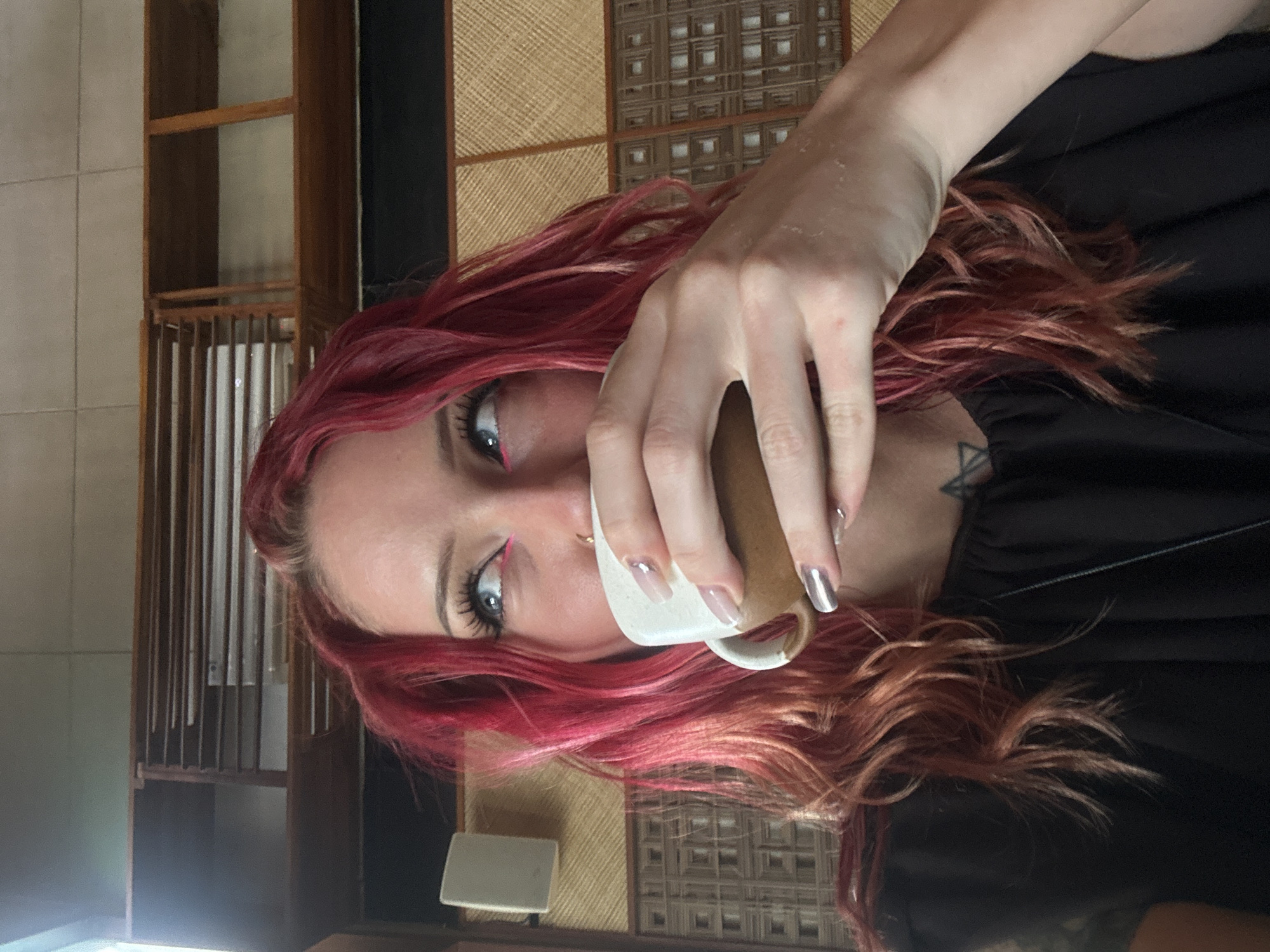 A woman with wavy red hair and blue eyes, wearing a black top, is drinking from a brown and white mug. Her left hand is holding the mug near her face. She is indoors, with a tatami mat and a dark shelf behind her. The location is North Kuta, Indonesia.