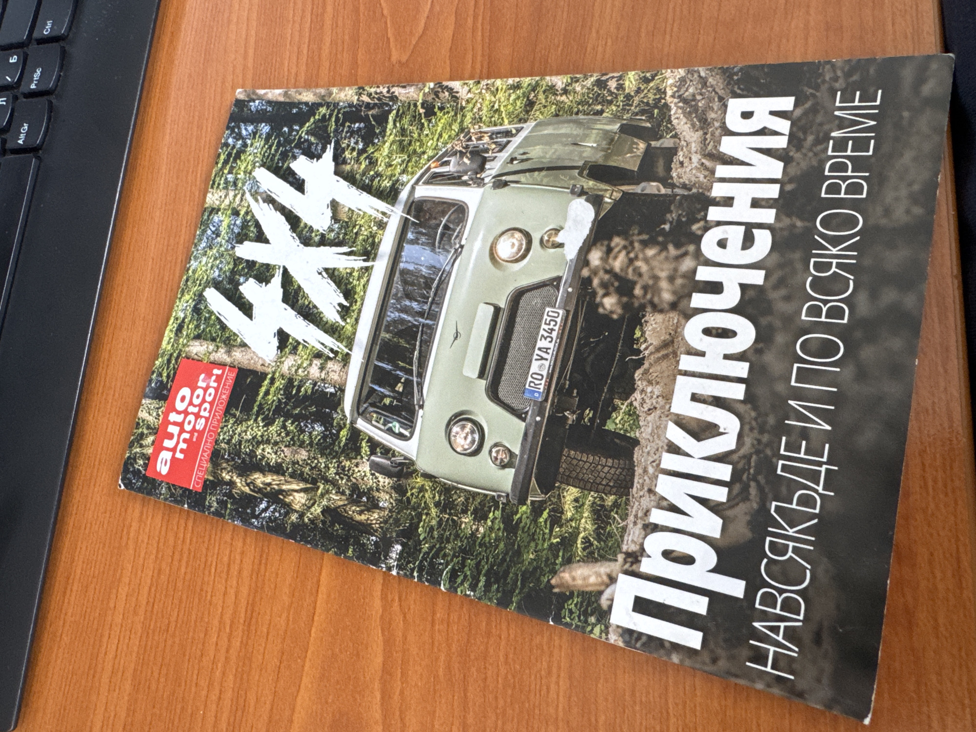 A magazine cover featuring an off-road vehicle is on a wooden surface, near a black keyboard. The magazine has the words "4x4" in large white letters on the upper left, with the words "auto motor sport СПЕЦИАЛНО ПРИЛОЖЕНИЕ" in red and white below. The vehicle in the image is an olive green, boxy off-road vehicle. The license plate on the vehicle reads "RO•YA 3450". Next to the vehicle are the Cyrillic words "Приключения" and "НАВСЯКЪДЕ И ПО ВСЯКО ВРЕМЕ" in white letters. There is no indication in the photo that it was taken in Sofia, Bulgaria.