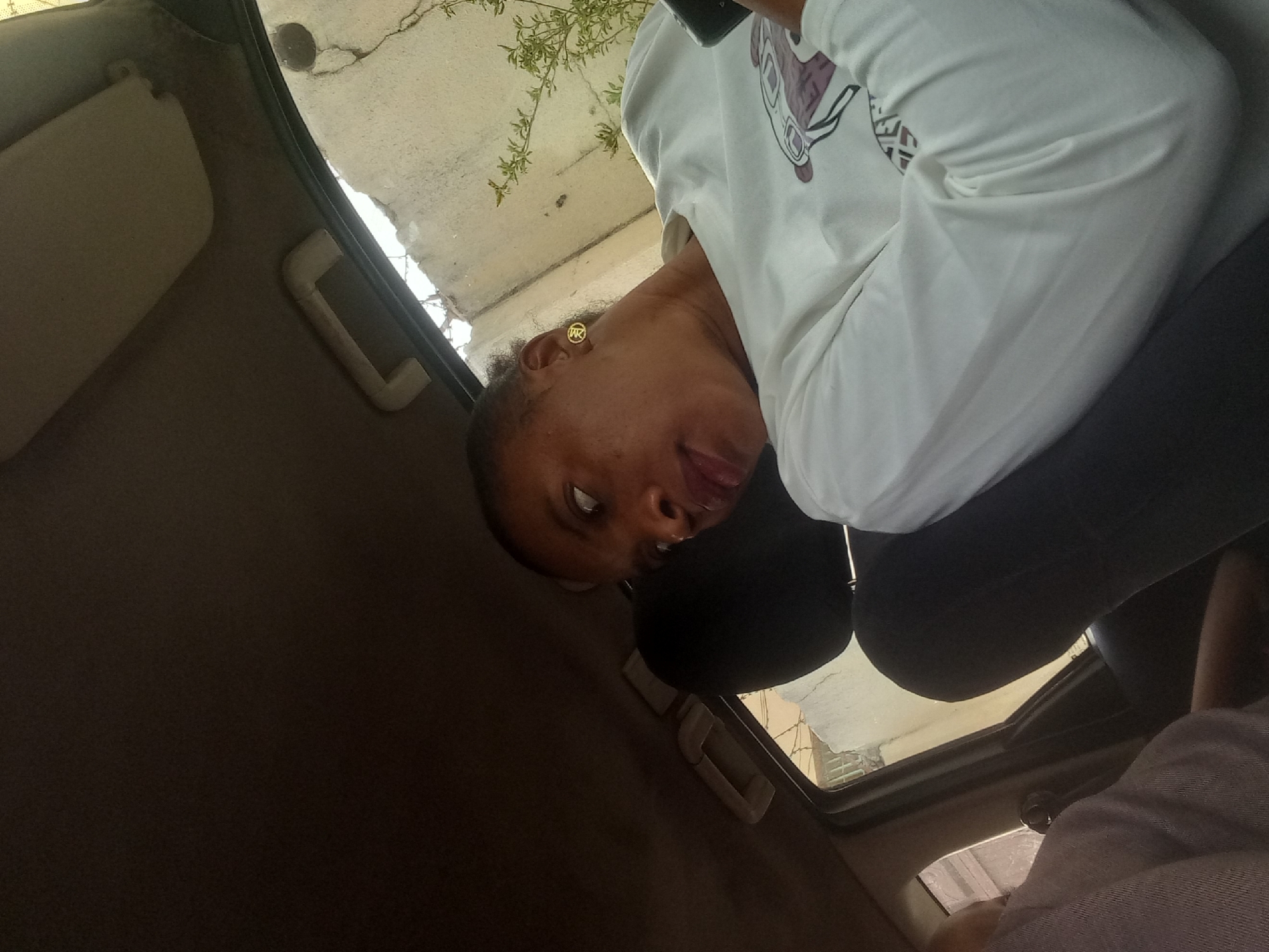The image captures a person inside a vehicle, likely in Barden Yana, Nigeria, during daylight hours. The photo is taken from a low angle, tilted, and looks upwards towards the subject.
The main subject is a person with dark skin, visible from the upper body upwards. They are wearing a white long-sleeved shirt featuring an abstract graphic design with purple/brown shapes. In their visible ear, they wear a gold-colored stud earring embossed with the letters "MK". Their dark hair is pulled back or styled close to their head. The person is looking directly at the camera with wide eyes and their tongue slightly out, suggesting a playful or casual expression. They are also wearing dark trousers or leggings. A dark object, possibly a mobile phone, is partially visible near the top of their head.
Inside the vehicle, parts of the car's interior are visible. A light-colored sun visor is seen towards the top left of the frame, and a beige grab handle is mounted near the window. The car's headliner and interior trim appear dark. Behind the subject's head, a dark headrest is visible. In the lower right corner, a glimpse of another person's limb, covered in light-colored, possibly striped fabric, suggests the presence of another occupant.
Outside the vehicle, through the window, a cracked concrete or paved surface is visible. Sparse green foliage, resembling a small plant or vine, grows on or near the concrete structure. The bright lighting indicates it is daytime, and the weather appears clear. The car seems to be stationary.
The overall impression is candid and lighthearted. The only visible text is the "MK" on the earring.