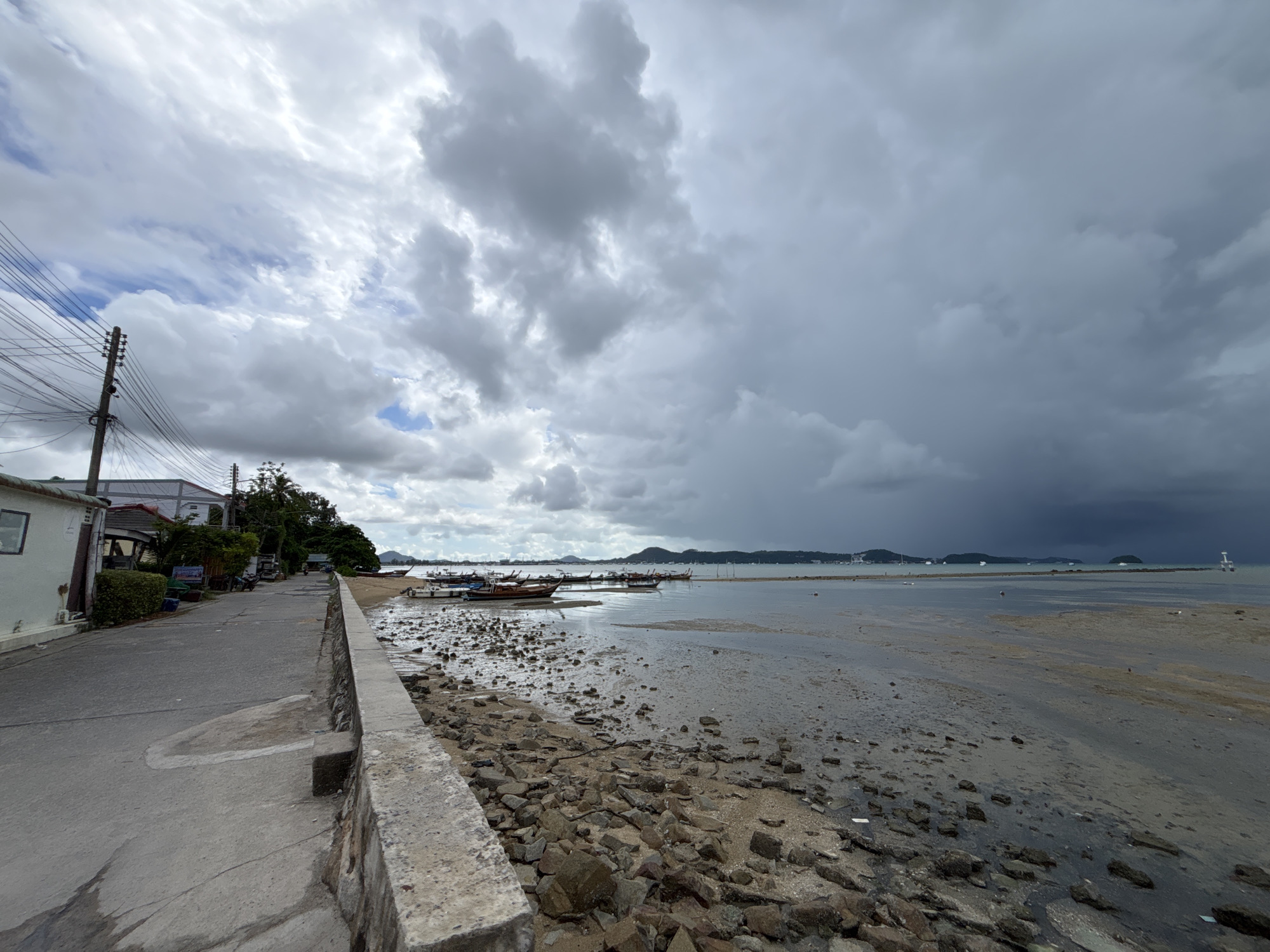 This is a wide-angle, eye-level, outdoor photograph taken in Rawai, Thailand. The image depicts a coastal scene with a paved road running along the left side, bordered by a stone wall. Several traditional wooden boats are moored in the shallow water to the right of the road. The sky is heavily overcast with dark, dramatic storm clouds dominating the upper portion of the frame, contrasting with lighter clouds and patches of blue sky towards the left. The ground near the water's edge is rocky and appears to be exposed at low tide. In the distance, a few more boats and structures are visible on the horizon. The overall atmosphere is moody due to the impending storm.