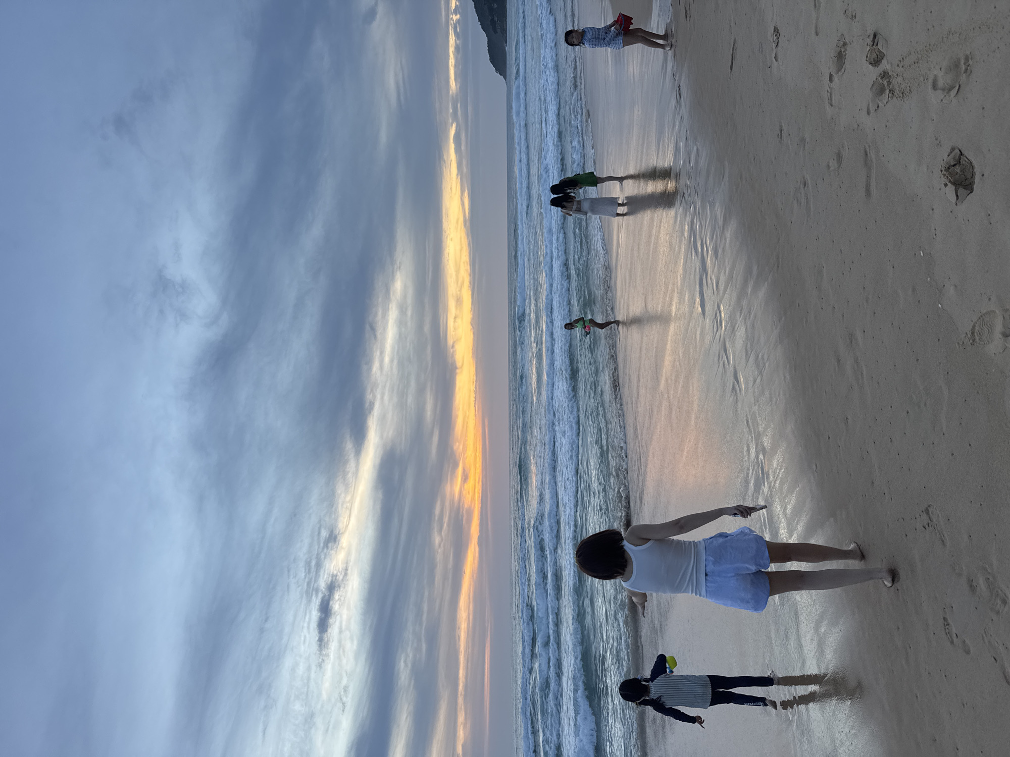 At dusk on Karon Beach in Karon, Thailand, several people are walking along the wet sand near the shoreline. The sky is filled with dramatic clouds tinged with orange and yellow from the setting sun, and the ocean is active with rolling waves. In the foreground, a young person in a striped long-sleeved swimsuit and dark pants walks to the left, carrying a yellow toy. A taller person in a white tank top and light blue shorts walks away from the camera towards the water, their reflection visible on the wet sand. Further out, near the water's edge, two people in light-colored dresses stand together, and another person in a green outfit is visible. A young person in a striped swimsuit and red bucket stands on the right side of the frame. The wet sand is marked with footprints.