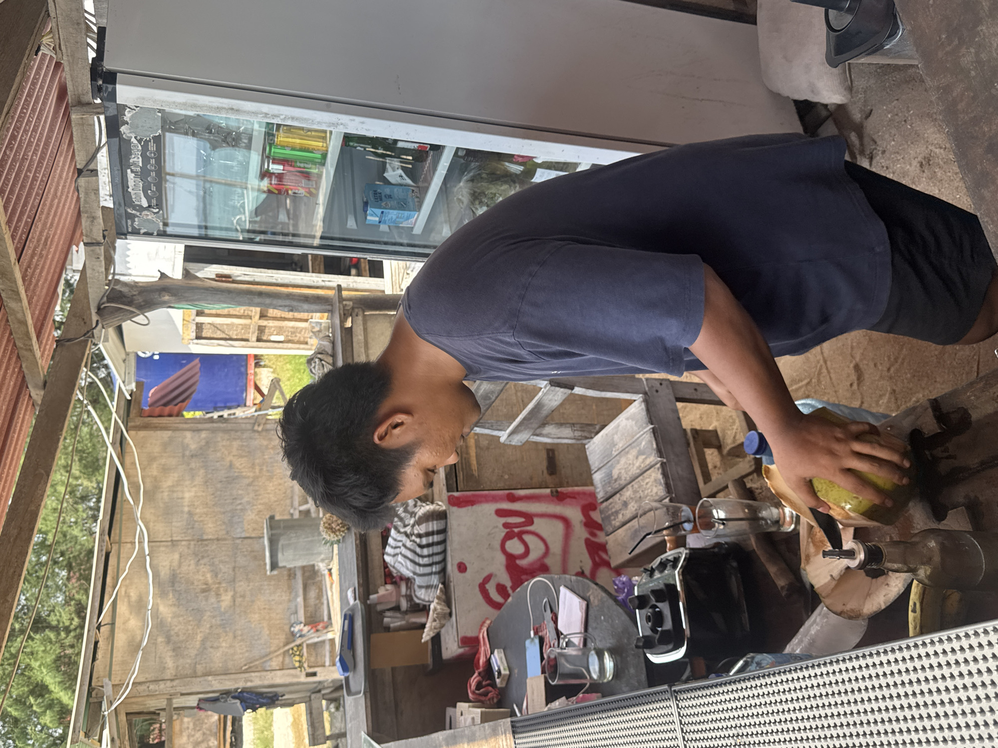 A man is shown preparing a coconut in what appears to be an outdoor food stall or small eatery in an unknown location in Indonesia. The man, wearing a dark blue t-shirt and black shorts, is holding a large knife and is in the process of cutting open a green coconut. The coconut is resting on a wooden surface, likely a table, alongside other items such as a blender, glasses, and bottles. The background reveals a rustic, open-air structure with wooden walls and a corrugated metal roof. A white refrigerator is visible to the right, stocked with drinks and what appears to be packaged food. Various shelves and storage areas are present, suggesting a functional, albeit simple, commercial setup. The environment appears to be somewhat unkempt, with debris on the ground and a casual, utilitarian atmosphere. The overall scene depicts a common street-side food vendor preparing fresh produce.