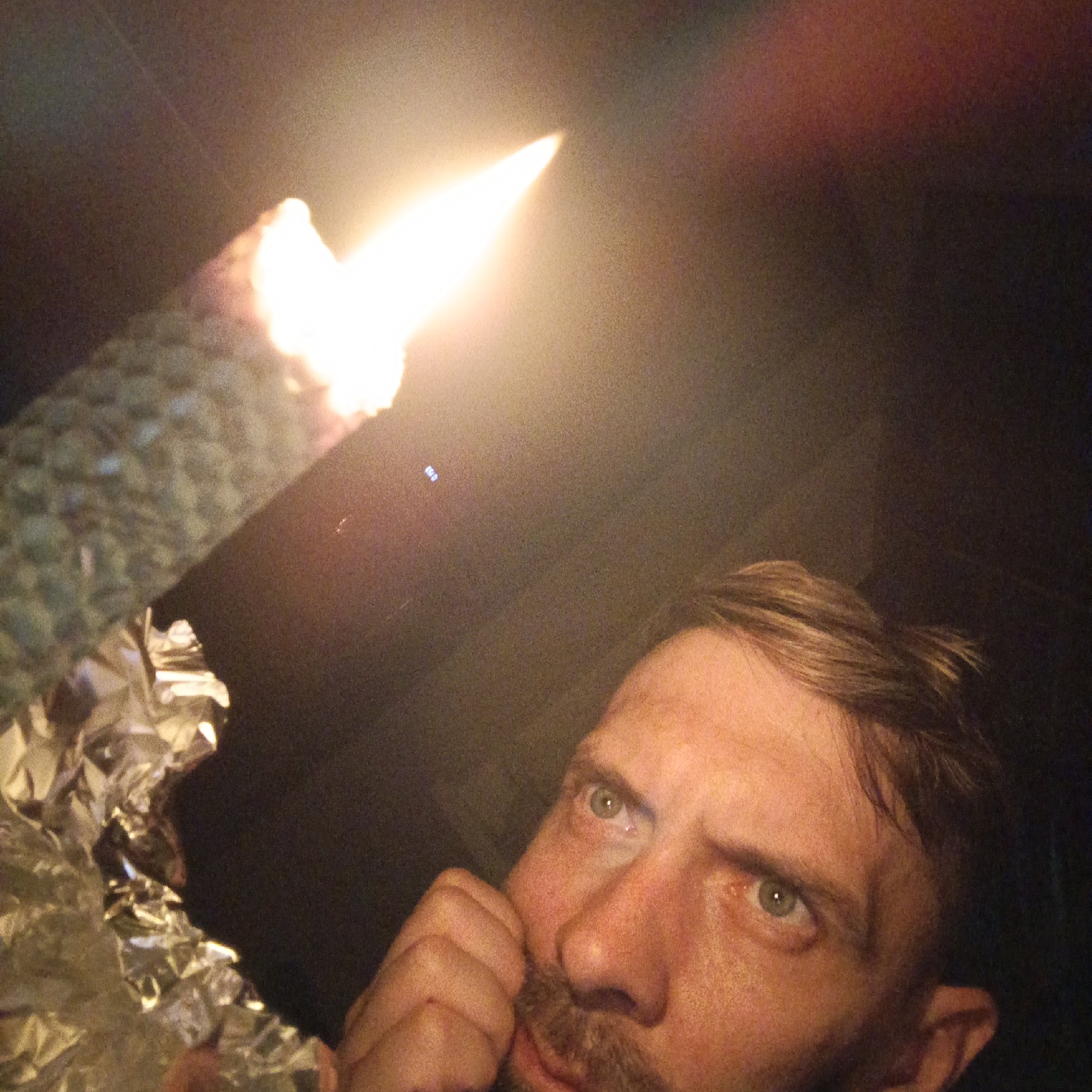A low-angle shot depicts a man with light brown hair, light complexion, and a goatee, looking up with a contemplative expression. His right hand is raised to his face, with his index finger near his cheek. To the upper left is a burning torch, possibly a makeshift one, wrapped in what appears to be aluminum foil. The light from the torch illuminates the man's face and creates a bright glare. The background is dark and indistinct, suggesting an indoor setting at night. There is no evidence in the image to confirm the location as Марковский, Russia.