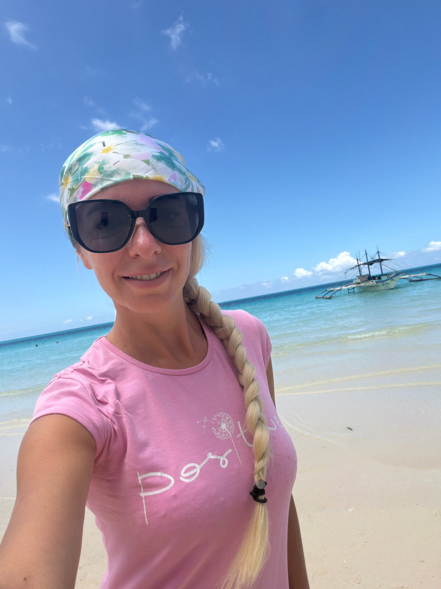 A blonde woman wearing a pink t-shirt and a floral headscarf takes a selfie on a white sand beach. She is wearing large black sunglasses and her hair is in a braid. In the background, the turquoise ocean stretches to the horizon under a bright blue sky with scattered clouds. A traditional Filipino outrigger boat, known as a bangka, is visible anchored in the shallow water. The scene depicts a sunny day at a tropical beach, likely in Malay, Philippines, given the presence of a bangka boat and the characteristic clear blue waters and white sand.