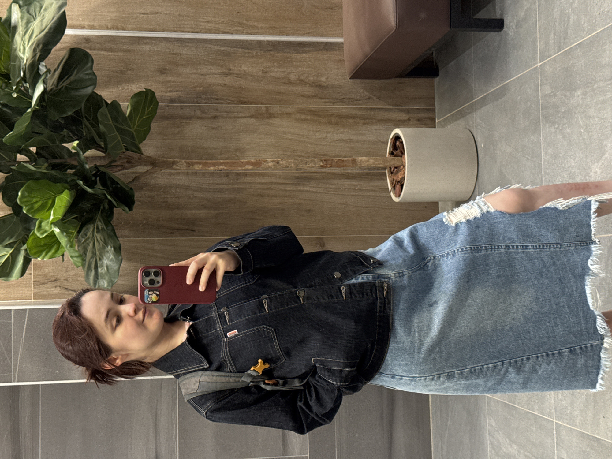 A young woman is taking a mirror selfie in what appears to be a modern restroom or fitting room. She is wearing a dark denim jacket and a light blue denim skirt with distressed detailing and a frayed hem. A grey backpack is slung over her left shoulder, with a bright yellow buckle visible. She is holding a red iPhone in her right hand, which is angled to capture the reflection. Her hair is pulled back into a messy ponytail, and she has a light makeup application.
The background features a juxtaposition of materials. To her left, a wall is tiled with large, rectangular grey tiles. To her right, a wall is paneled with vertical, wood-grain textured tiles. A tall potted plant with large, dark green leaves stands in a light grey cylindrical pot between the woman and the wood-paneled wall. Further to the right, a dark brown, upholstered bench with black metal legs is visible. The floor is tiled with large, square grey tiles, and grout lines are visible, suggesting a clean, contemporary aesthetic. There is no direct indication of the location being in Izhevsk, Russia, or any specific city.