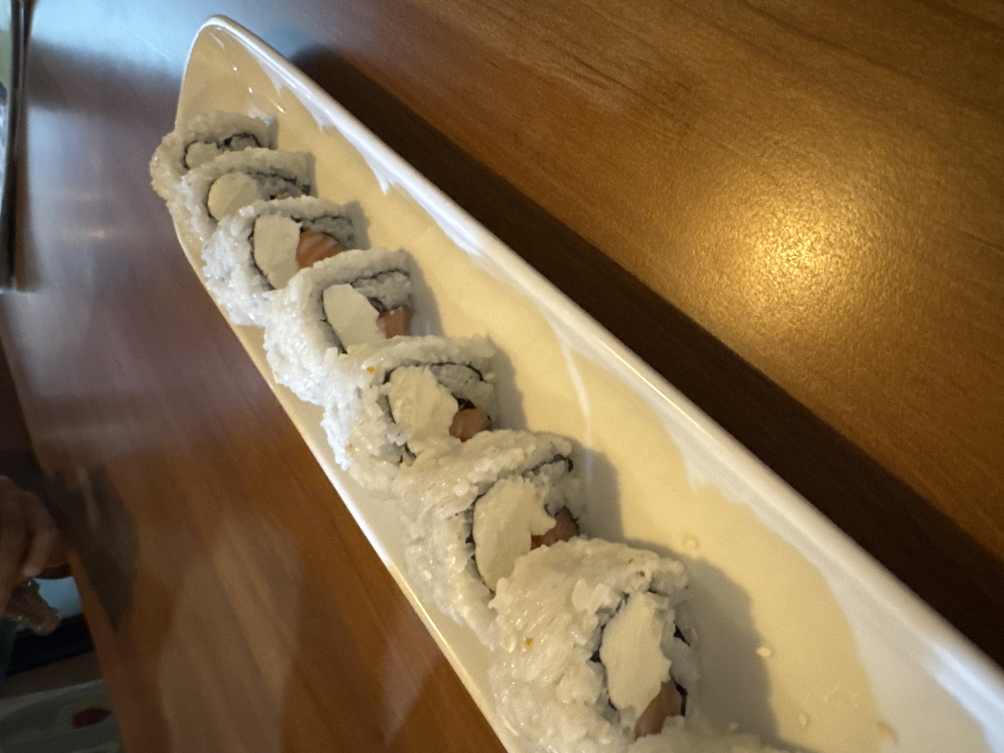 This close-up image features a long, white rectangular plate holding a row of sushi rolls, positioned on a warm, medium-brown wooden table. There are at least eight pieces of uramaki-style sushi visible, each generously covered in white rice. The fillings include light-colored fish, possibly salmon, and a prominent white, creamy ingredient, likely cream cheese. A hand is partially visible in the bottom left corner, suggesting the act of dining or serving. The setting is an indoor dining area, consistent with a meal being presented or consumed. The general atmosphere is casual, and the provided context indicates the location is Kuta Selatan, Indonesia. No specific time of day or weather is evident, nor are any emotions discernible or text visible.