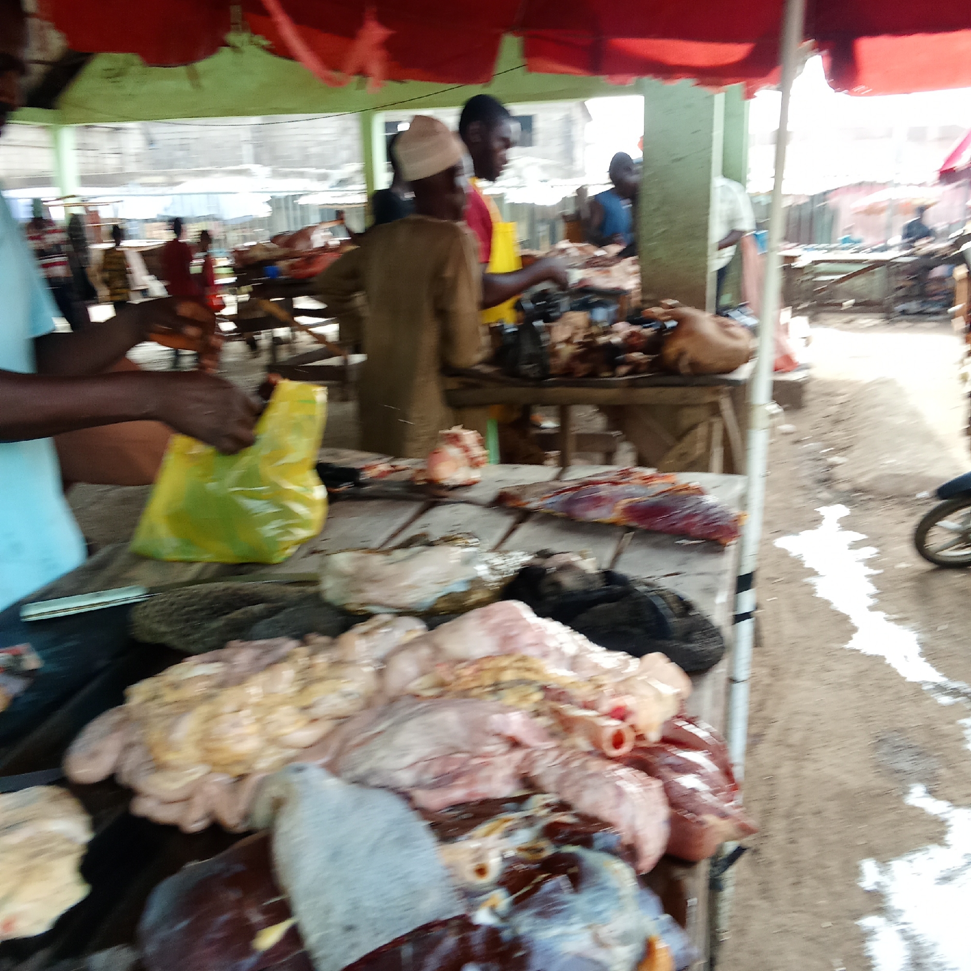 This image captures a bustling meat market scene, likely in Akwanga, Nigeria, during daylight hours. The primary focus is on a wooden stall displaying a large quantity of raw meat, including various cuts of muscle tissue, intestines, and organs such as liver, some still with fat attached. The meat is laid out on what appears to be a light-colored surface or cloth covering the wooden table.
In the foreground on the left, a person with dark skin, wearing a light blue short-sleeved shirt, is actively placing items into a bright yellow plastic bag. A knife or similar tool lies on the table next to their hands.
Behind this central activity, several other individuals are visible. One person in a light brown top and a light-colored cap stands behind another meat stall, while further back, a man in a red shirt and yellow apron is also tending to meat. Other figures are present in the midground and background, suggesting a busy environment. The market is covered by red and light green awnings or tarps, providing shelter from the sun.
The ground beneath the stalls is dirt, with patches of white residue, possibly lime or ash, scattered on it. In the far right background, a partial view of a motorcycle wheel is visible, parked on the dirt path. The overall atmosphere is one of an active, open-air market dedicated to selling fresh meat. There is no visible text in the image.