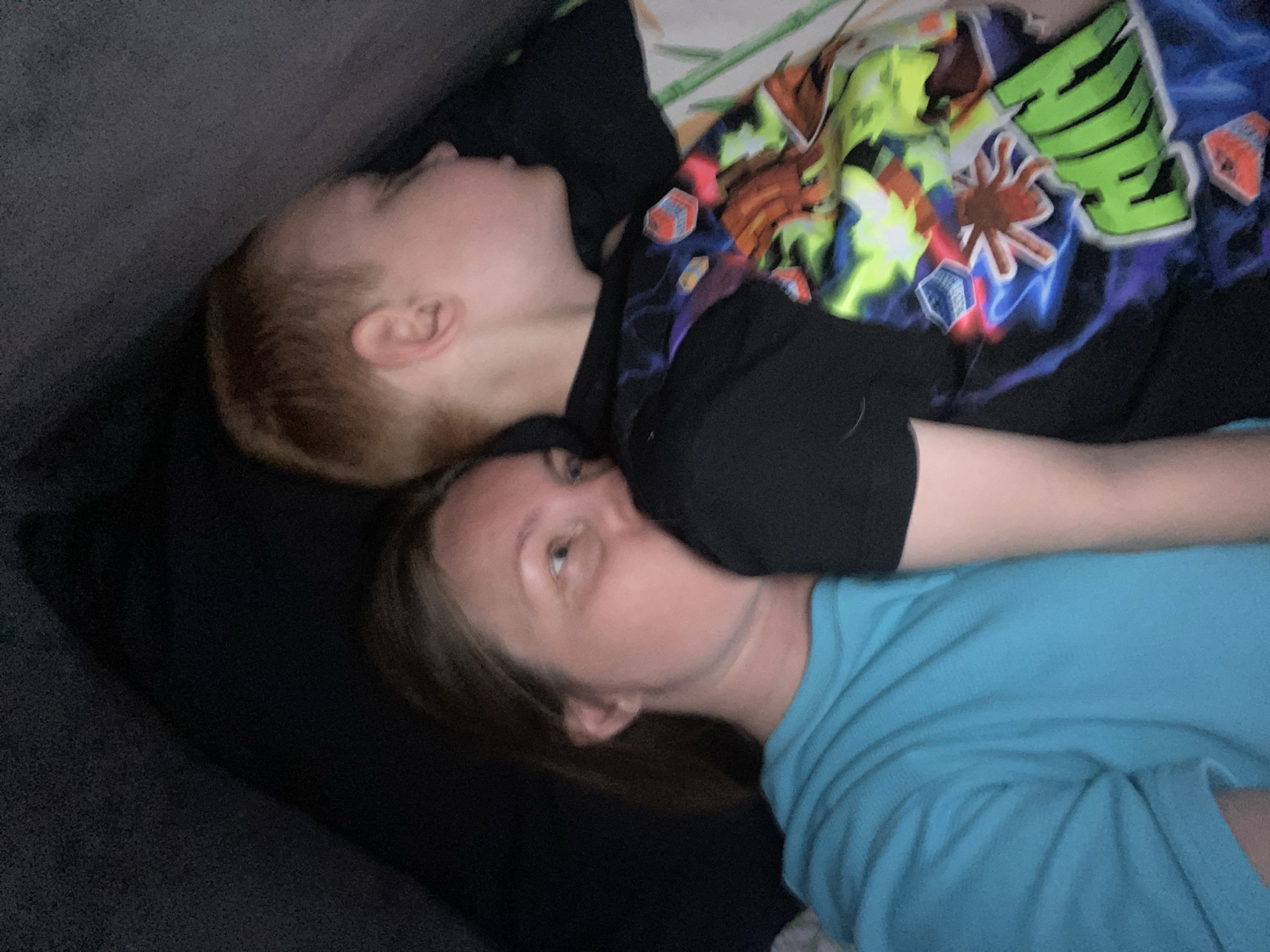 Two people, likely a mother and child, are lying down. The child, with red hair, is wearing a black shirt with colorful designs. The adult is wearing a light blue shirt. They are lying on what appears to be a dark-colored couch or cushion. No information suggests this image was taken in Noviy, Russia.