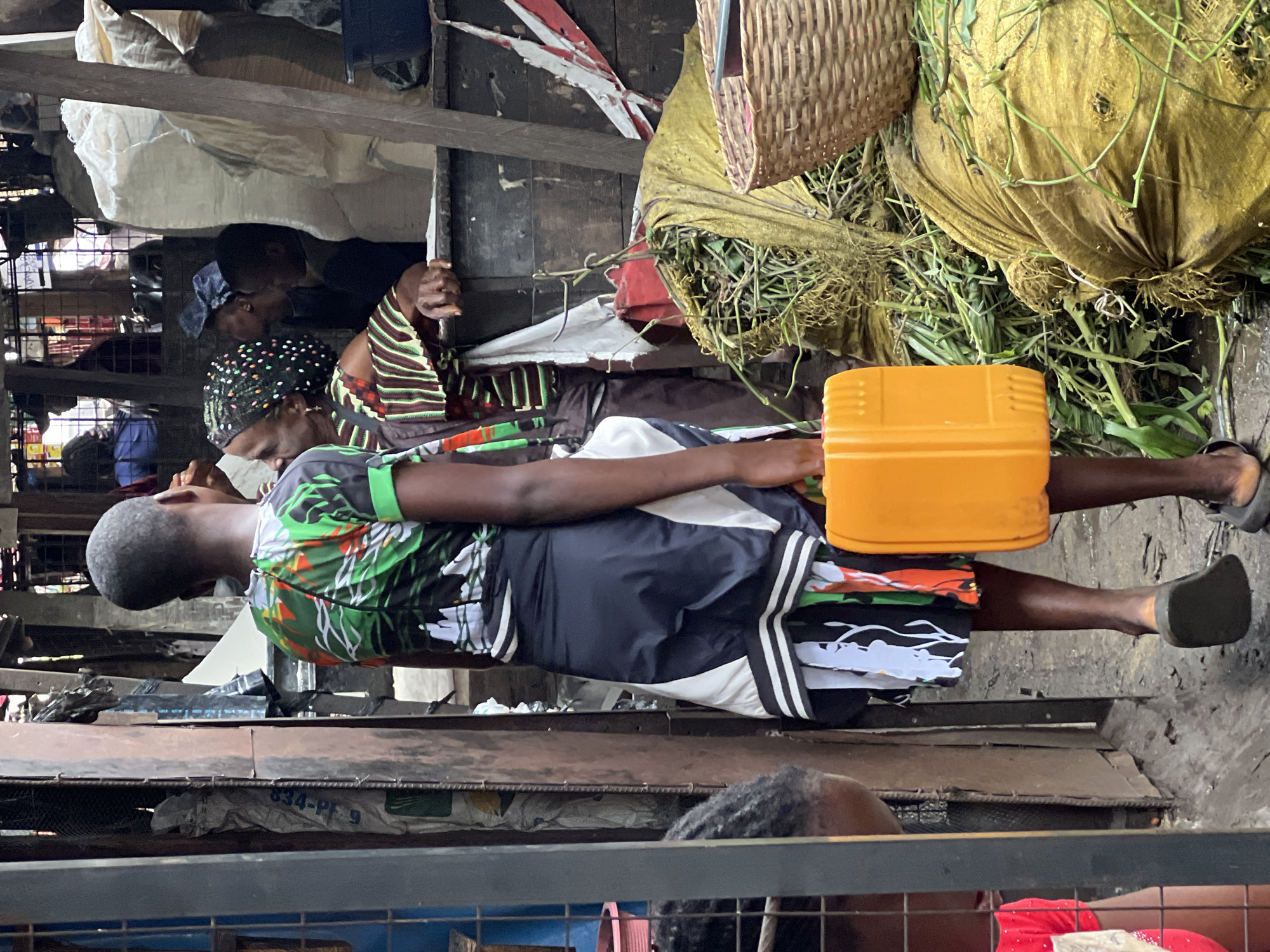 A street market scene in Nkpor, Nigeria, is depicted from a medium shot, focusing on two women and a child. The woman in the foreground, a young girl, walks away from the camera, wearing a vibrant green, black, and white patterned short-sleeved shirt and a navy skirt over a patterned underskirt. She carries a bright orange plastic container in her right hand. To her left, another woman, older, faces the girl and is dressed in a brown and green striped top with a patterned headscarf. Behind them, other individuals and market stalls are visible, with sacks of produce and woven baskets scattered around. The ground appears to be damp and uneven, suggesting an outdoor market environment.