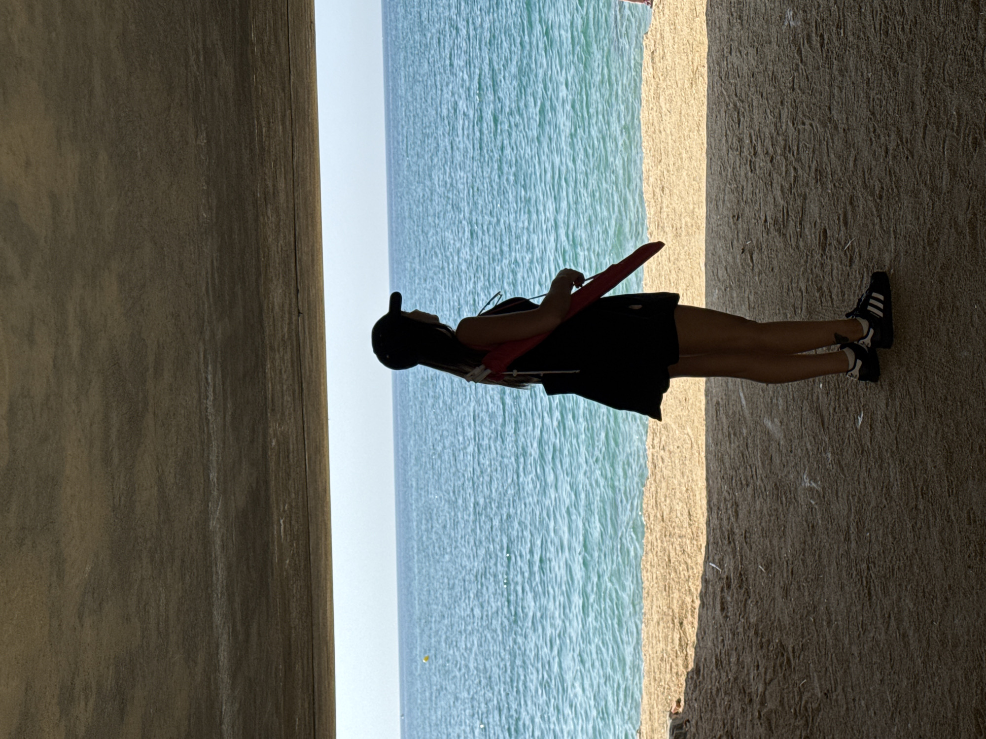 A silhouetted woman stands on a sandy beach, facing a bright blue ocean. She is wearing a black baseball cap, a black top, and black shorts. A red object, possibly an umbrella or a poster tube, is tucked under her right arm. Her left foot is slightly in front of her right, and she has black athletic shoes with white stripes. The woman is standing under a concrete overhang, which casts a shadow over the foreground. The sand is light brown and textured, with some small debris scattered on it. The ocean water is calm with gentle ripples, and the horizon line is clear against a light blue sky. A small yellow buoy is visible in the water to the left. The scene is captured from a medium-low angle, looking towards the sea. The location is identified as a beach in Arenys de Mar, Spain.