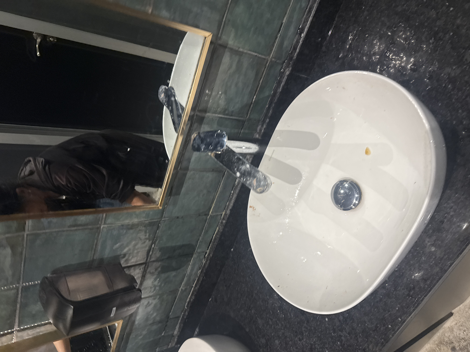 The image shows a bathroom sink with a white, oval-shaped basin. A chrome faucet is mounted above the sink. The countertop is black with what appears to be a glossy finish. There is a mirror in the scene, reflecting the sink and tiles on the wall. The reflection shows the sink faucet and some other objects. The tiles appear to be grey and textured. The location in Plovdiv, Bulgaria, is not evident from this image.