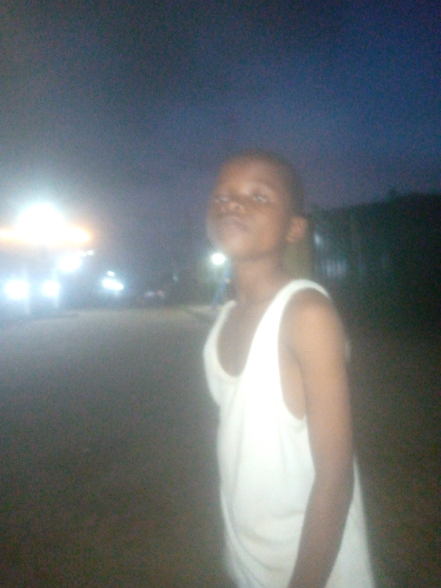 A young person stands on a dimly lit street at night. The person is wearing a white tank top. In the background, there are streetlights and the headlights of cars. The location appears to be a residential area with dark foliage visible. The image was taken in Ikorodu, Nigeria.