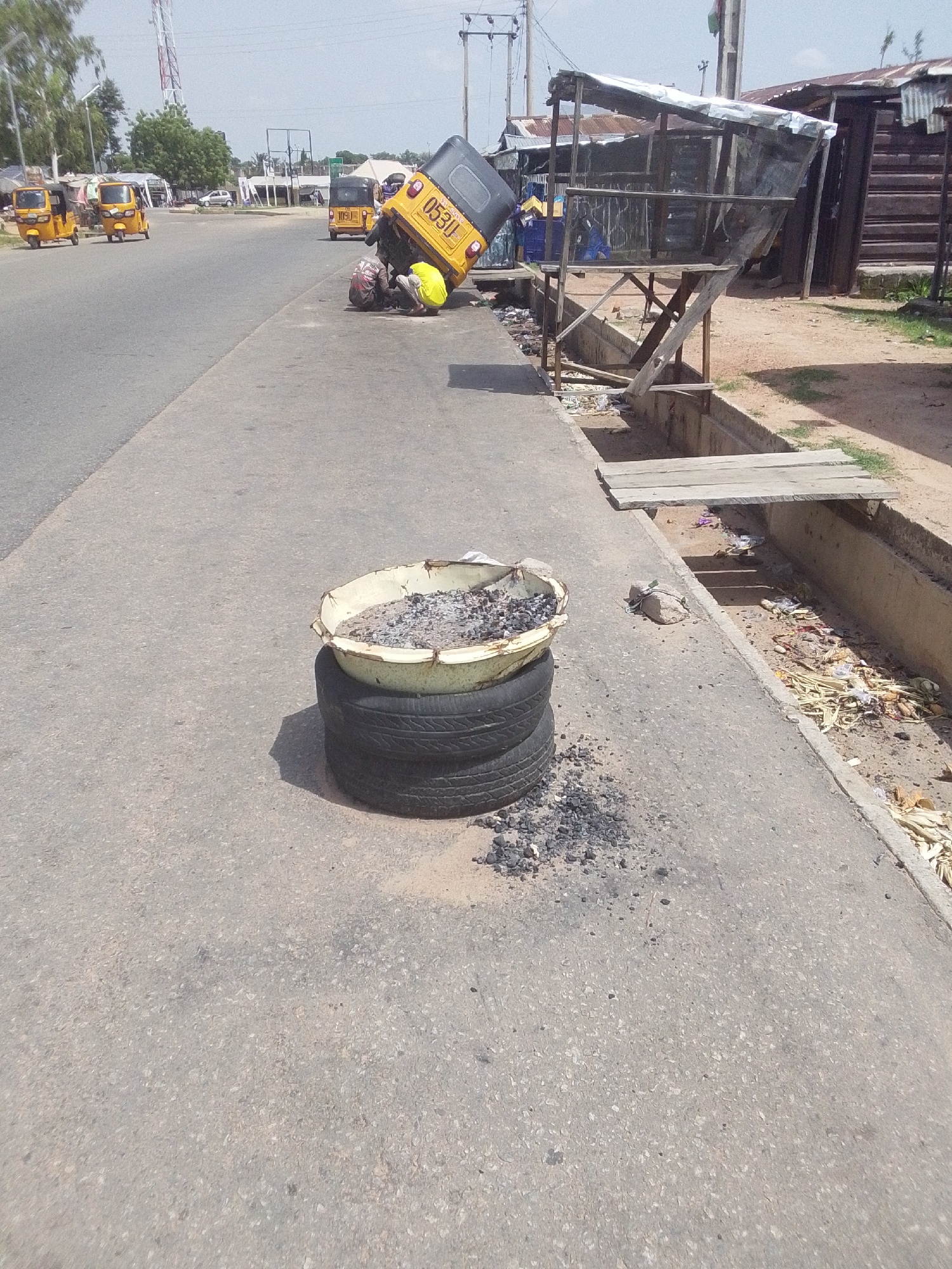 The image depicts a scene on a street in Jalingo, Nigeria. In the foreground, a makeshift brazier is constructed from two stacked tires with a large, shallow metal basin filled with ash and embers placed on top. The brazier is situated on an asphalt road. To the left of the brazier, the road curves away.
In the middle ground, a yellow motorized tricycle, commonly known as an "okada" or "tuk-tuk," has overturned, lying on its side. Several people are gathered around the overturned tricycle, some appearing to be assisting or inspecting it. One individual is wearing a bright yellow garment, while others are dressed in darker clothing. The tricycle's license plate is visible, with the characters "05311" clearly discernible.
Further in the background, more yellow tricycles are parked or moving along the road. On the right side of the road, there are several makeshift stalls with corrugated metal roofs, typical of a market or roadside vendor area. A concrete drainage ditch runs alongside the road, filled with debris and some discarded items. A simple wooden plank spans a section of the ditch, serving as a makeshift bridge.
The overall scene suggests a daytime setting under clear skies, with bright sunlight casting shadows. The environment appears to be a busy roadside area in an urban or semi-urban Nigerian location. The overturned tricycle and the activity around it might indicate a minor accident or a mechanical issue. The presence of the brazier suggests possible street food vending or some form of outdoor cooking.