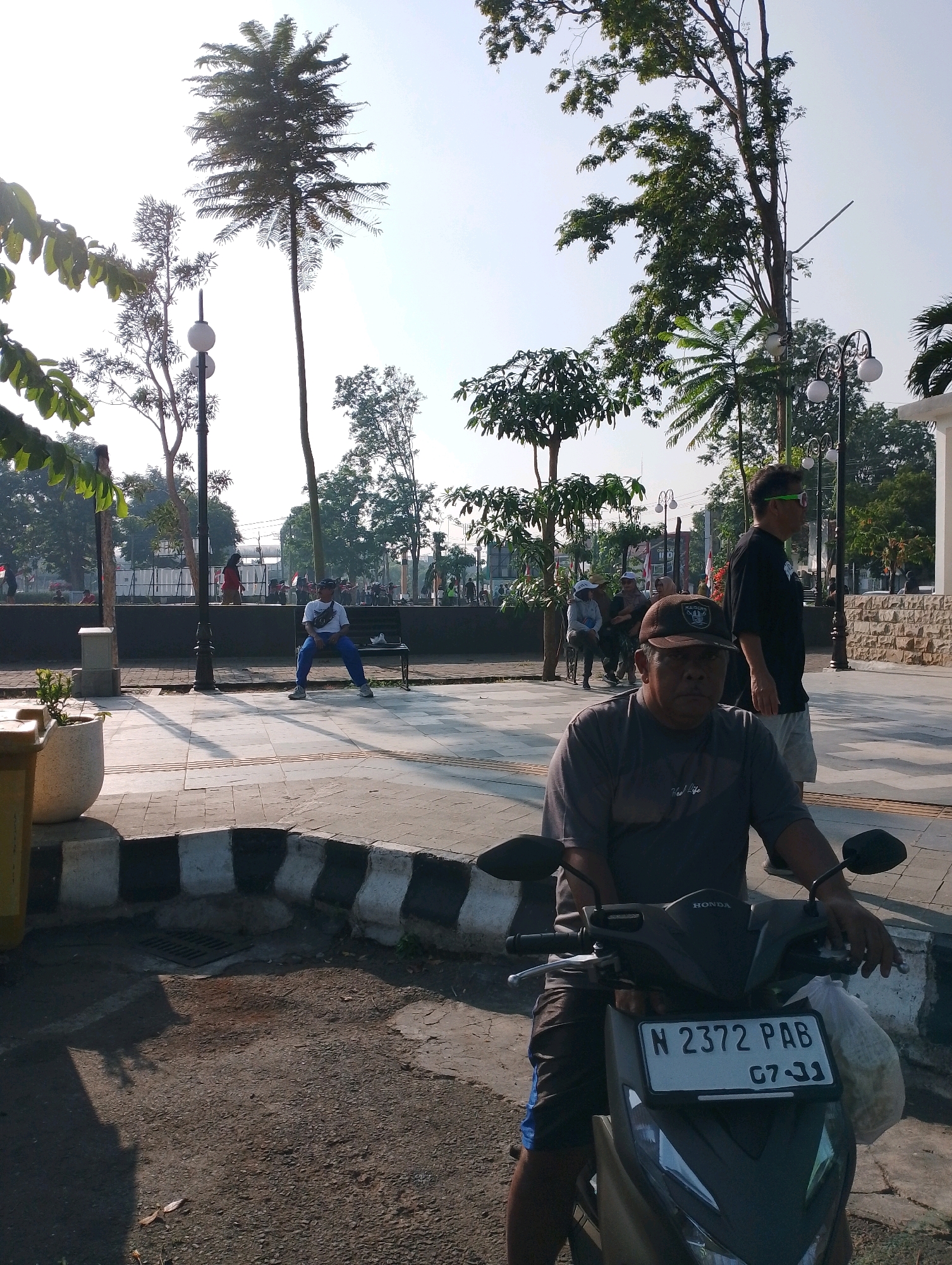 An outdoor scene in Probolinggo, Indonesia, captures a man on a scooter in the foreground, facing forward and looking at the camera. He wears a dark gray t-shirt, a brown baseball cap, and blue shorts. His hands are on the scooter's handlebars. The scooter's license plate reads "N 2372 PAB 07-21". Behind him, a man in a black t-shirt and light shorts stands facing left, wearing green-tinted glasses. Further in the background, a person sits on a park bench in a white t-shirt and blue pants, with their legs spread apart. The setting includes trees, lampposts, and paved walkways, suggesting a public park or plaza. In the distance, other people can be seen walking and standing, and there are various structures, possibly buildings or fences. The lighting indicates it is daytime, with sunlight casting shadows on the ground.