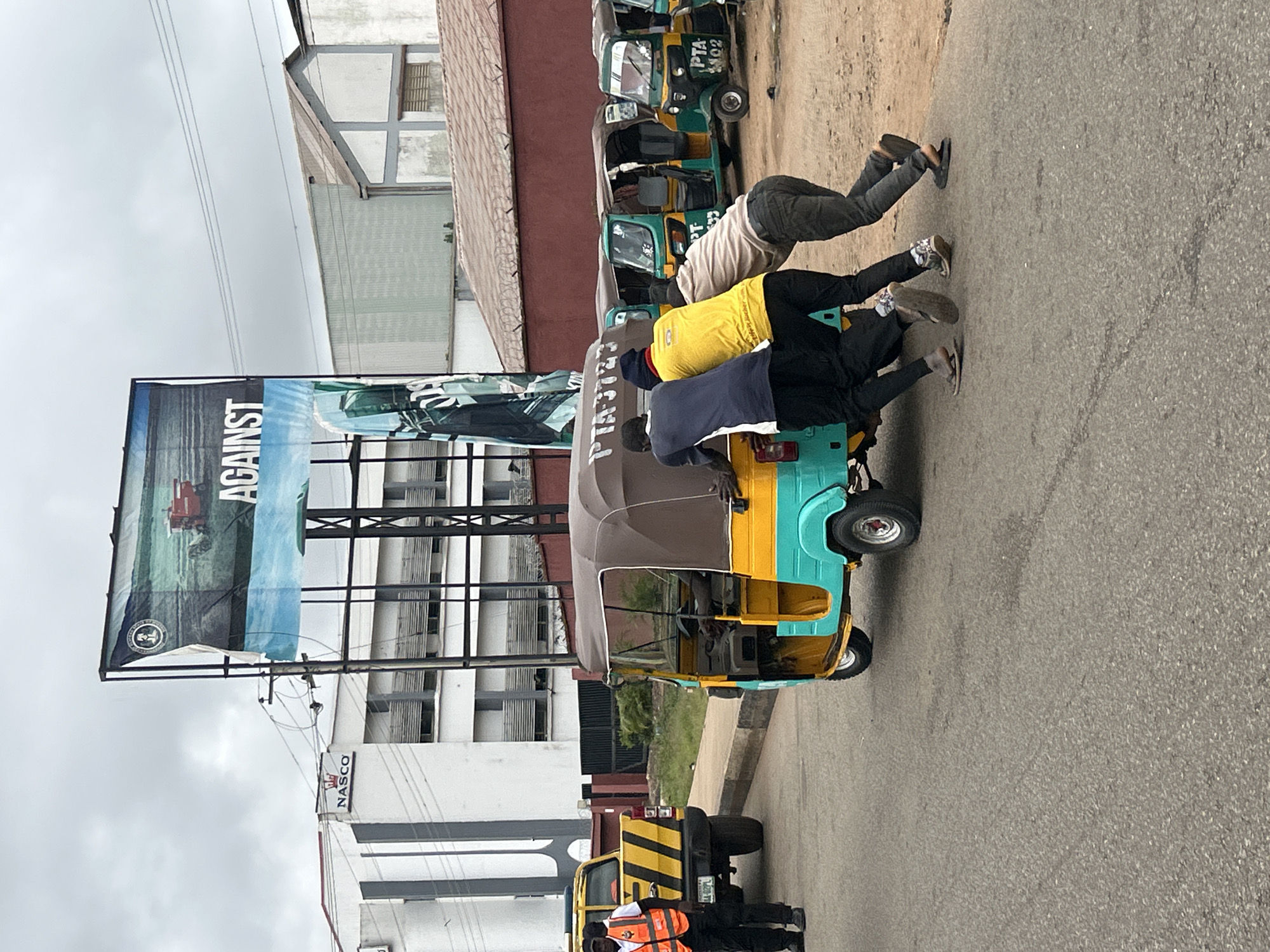 In the city of Jos, Nigeria, on an overcast day, a teal and yellow auto-rickshaw is being pushed by four men. The auto-rickshaw is in the foreground, angled to the right. Three men are visible pushing the back of the vehicle, with their bodies leaning into the effort. Their legs are bent, and their feet are on the asphalt road. One man in a dark blue shirt is furthest left, followed by a man in a bright yellow shirt, and then a man in a light brown shirt on the right. Another man, wearing a dark blue beanie and a dark blue shirt, is partially obscured behind the first man.
To the left of the auto-rickshaw, a yellow pickup truck with black and yellow stripes on its side is parked. A person in an orange safety vest stands to the left of the pickup truck, facing away from the viewer. The truck has a license plate that appears to read "AA-82PL."
In the background, there are several buildings. A white building with a dark gray facade and large windows is visible on the left. A billboard advertising "AGAINST" with an image of agricultural machinery is prominently displayed above the buildings, supported by metal poles. Further in the background, a long, low-rise building with a brown roof and light gray walls is visible. Another auto-rickshaw, painted green and yellow, is parked in the middle ground behind the auto-rickshaw being pushed, and additional auto-rickshaws are visible to its right. The road is paved with asphalt, and there is a curb to the right of the road. The sky is overcast, suggesting a cloudy day.