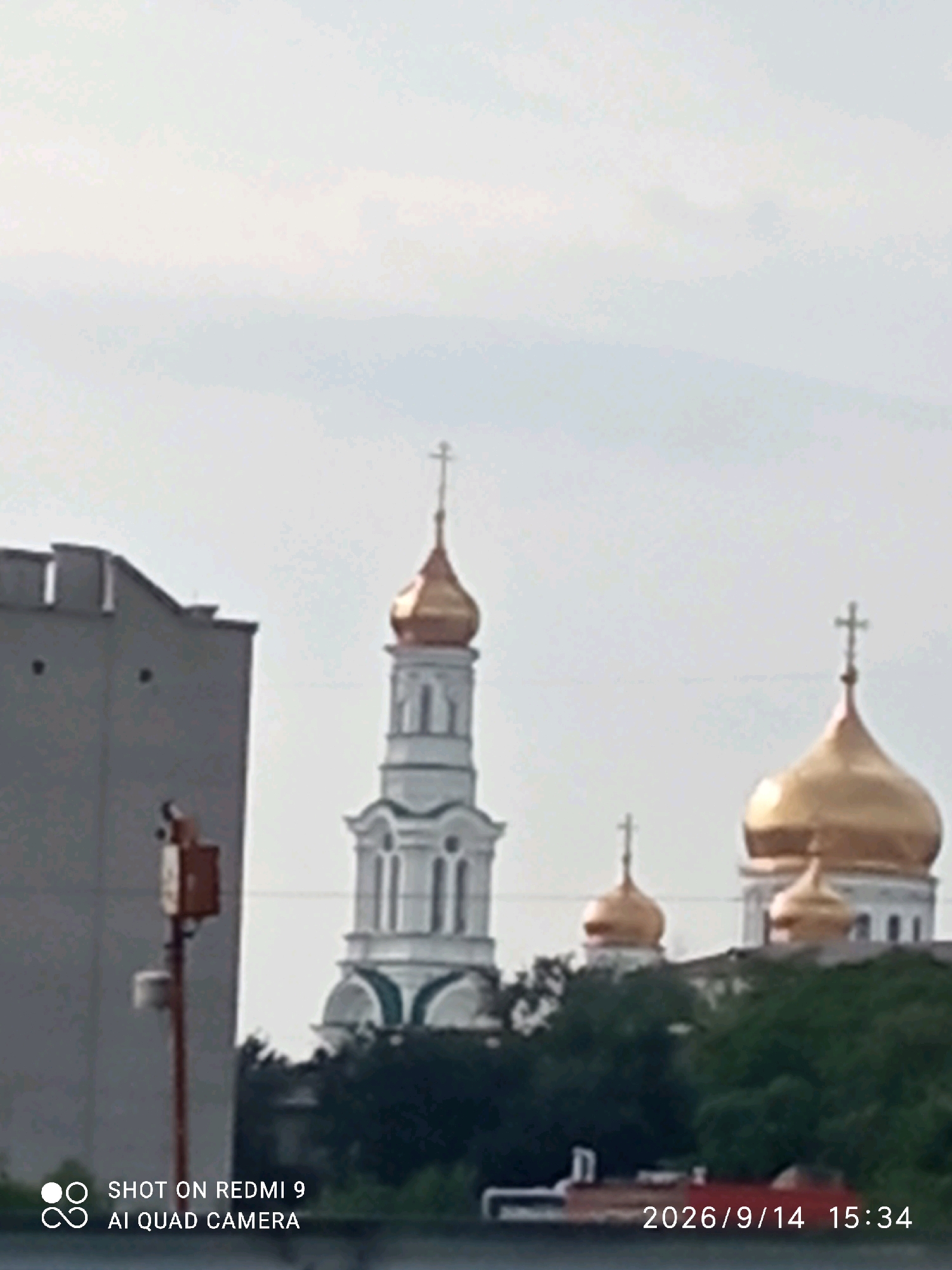 The image depicts a cityscape with prominent religious architecture under a cloudy sky. In the foreground, a gray, blocky structure with a flattened top is visible on the left. Attached to this structure is a pole with a small, orange box-like object at the top, possibly a camera or sensor. To the right of this structure, a white bell tower with a golden dome topped by a cross rises, featuring arched windows and decorative elements. Further to the right, partially obscured by trees, are additional golden domes, some larger and more bulbous, also topped with crosses. The scene is framed by lush green trees and foliage at the bottom. The lower right corner of the image displays a date and time stamp: "2026/9/14 15:34" and the text "SHOT ON REDMI 9 AI QUAD CAMERA" in the bottom left. This image appears to be taken from a distance, possibly from a building, looking across a developed area.