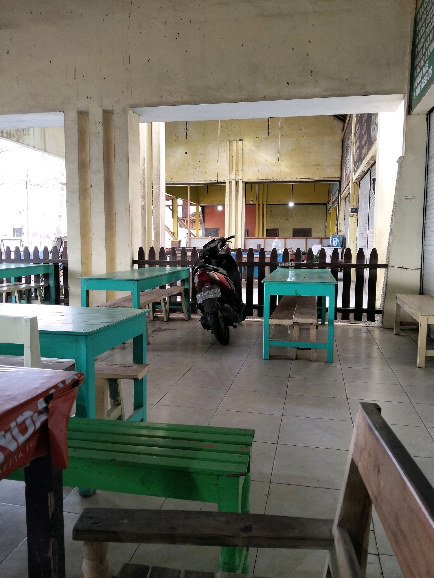 The image depicts an outdoor seating area of a food establishment located in Tasikmadu, Indonesia. The setting is an open-air pavilion with a tiled floor and a concrete ceiling supported by pillars. Several simple tables and benches are arranged for seating. The tables are painted in bright teal and green colors, with wooden benches accompanying some of them. One table is covered with an orange-and-white patterned cloth. In the background, a black motorcycle is parked. A wooden fence separates the seating area from the interior of the establishment, where glimpses of shops with closed shutters are visible, along with some signage. The overall atmosphere appears to be that of a casual, local eatery.