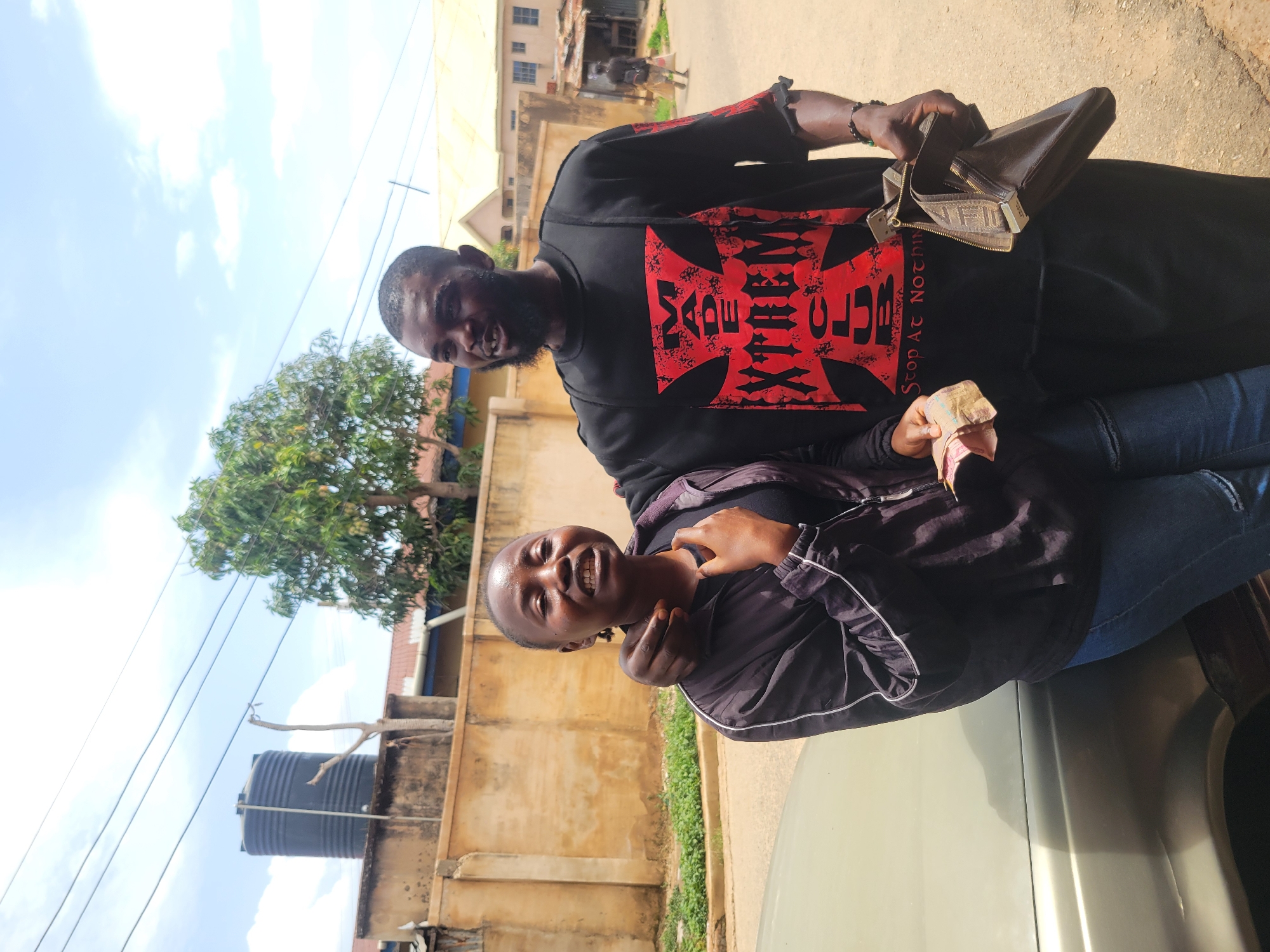 Two individuals are photographed outdoors in Jos, Nigeria. A dark-skinned man with a beard is wearing a black sweatshirt featuring a red graphic and holding a brown wallet and folded currency. A younger dark-skinned woman in a black jacket with white trim and blue jeans is smiling and touching her neck. They are standing near a light-colored vehicle, possibly a car. The background shows buildings, a water tank, vegetation, and a clear sky.