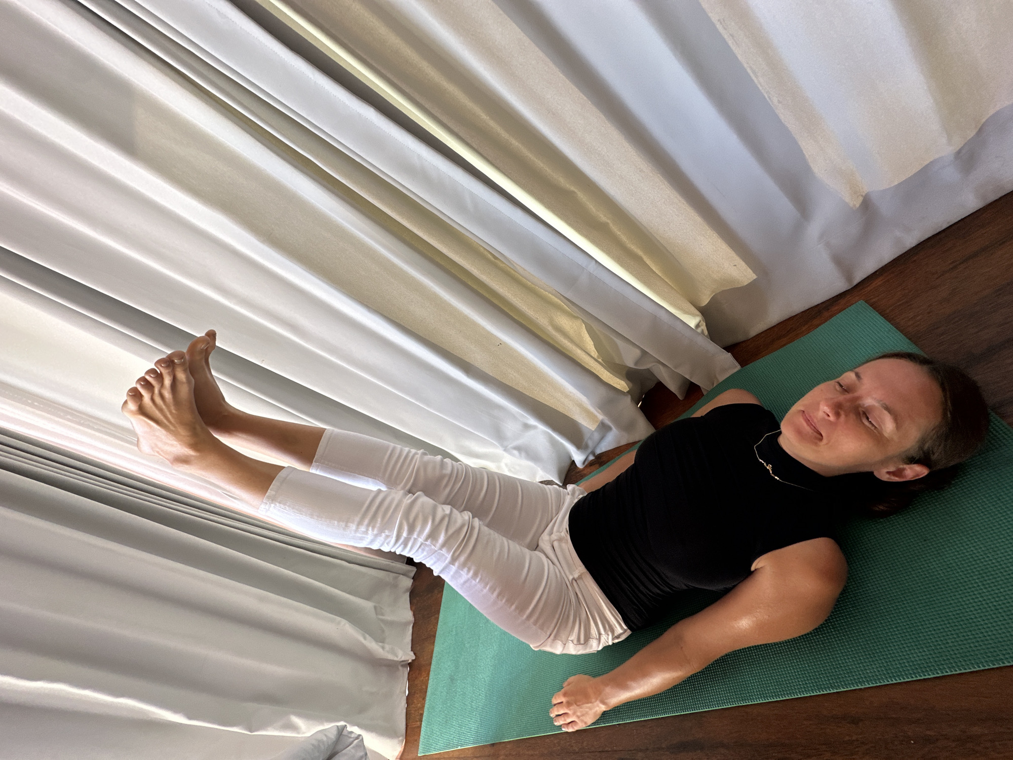 A woman is lying on her back on a green yoga mat on a wooden floor. She is wearing a black sleeveless top and white ruched leggings. Her legs are raised vertically in the air, with her feet pointed. Her eyes are closed, and her head is tilted back. She has a thin gold necklace. The background consists of light-colored curtains that are partially drawn. The scene appears to be an indoor yoga or meditation space, possibly in a resort or home in Nusa Penida, Indonesia.