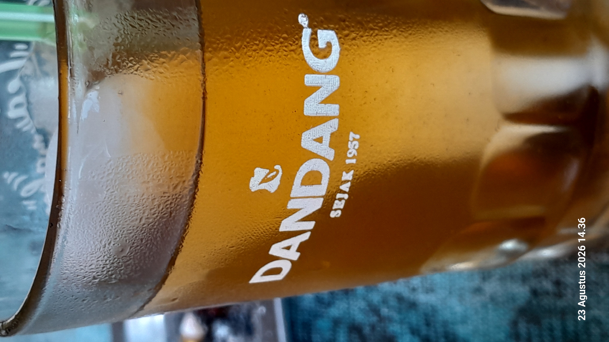 The image shows a close-up of a glass of iced tea with condensation on the outside. The brand "DANDANG" and "SEJAK 1957" are visible on the glass. A date stamp at the bottom of the image reads "23 Agustus 2026 14.36". The background is blurred and appears to be a teal-colored textured surface. There is no indication of the city of Gemaharjo, Indonesia, in the image.