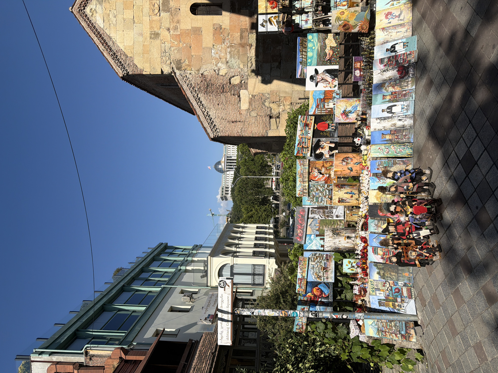 This image depicts a street scene in Tbilisi, Georgia, likely in the old town area, featuring an outdoor art and souvenir market. In the foreground, a collection of paintings and handcrafted dolls are displayed on a rack and along the cobblestone sidewalk. The paintings showcase various subjects, including cityscapes of Tbilisi, still lifes, and portraits. Several of the paintings are of buildings with distinct architectural features. Among the dolls, there are figures dressed in traditional attire.
To the left of the market, a multi-story building with a white facade and green trim is visible. Signage is present on a pole near the market, with text in Georgian and English, one of which reads "Ikhtang Gorgasali Square."
To the right, an old, weathered stone building with a brick roof and arched window stands, suggesting historical architecture. In the background, a large, modern building with a dome and flags is visible on a hilltop, possibly the Presidential Palace, and a construction crane further in the distance. The sky is clear and blue, indicating a sunny day. The environment appears to be an urban setting with a mix of old and new architectural styles.