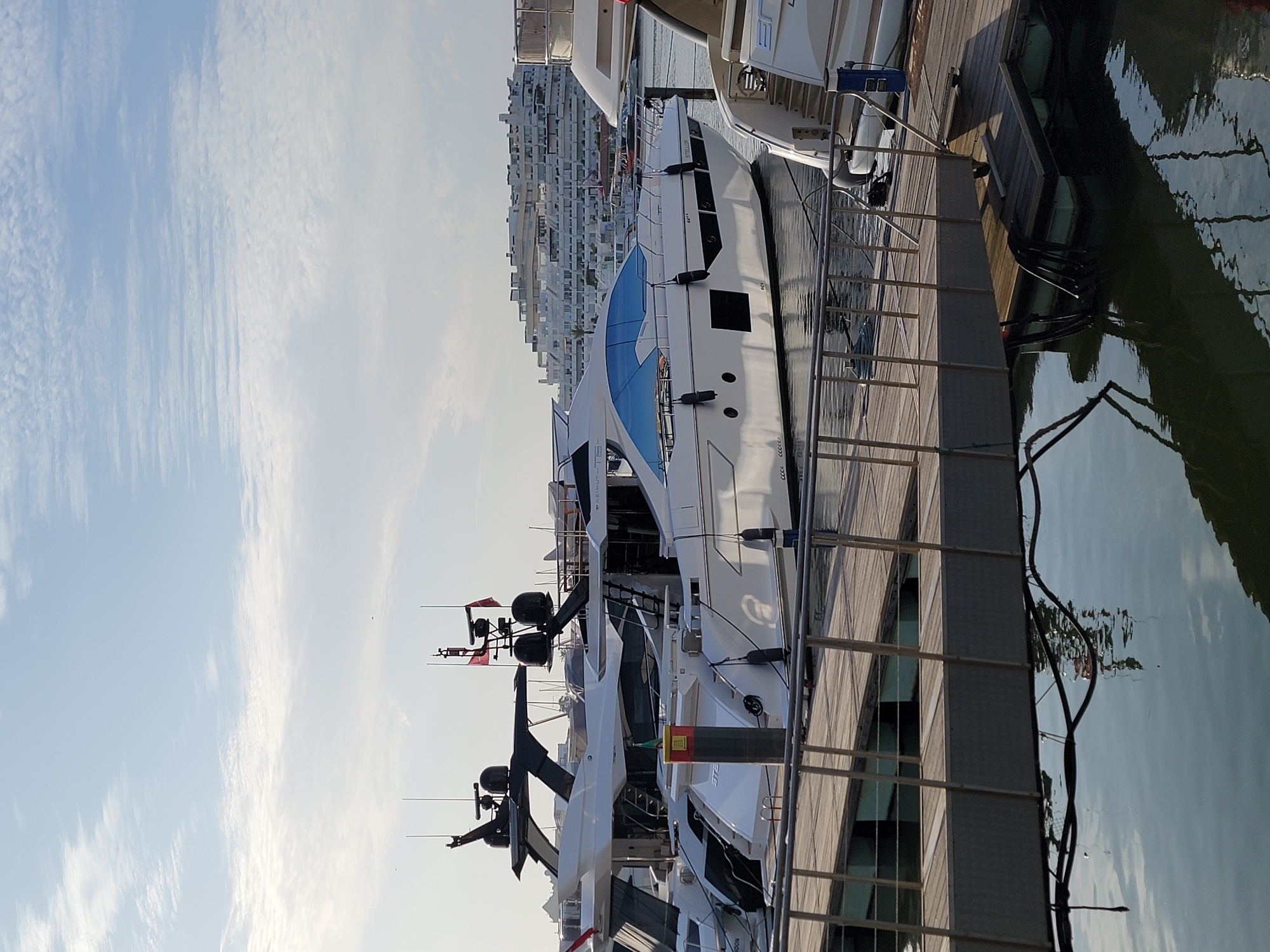 The image shows a waterfront scene in Quarteira, Portugal, likely in the late afternoon given the lighting. The primary subjects are several large, modern yachts docked along a wooden pier. These yachts are sleek and white, with large windows and various antennas and equipment on their decks. One yacht in the foreground has a distinctive blue-tinted glass section on its upper deck. The water beside the pier is calm and reflects the sky and the yachts.
In the background, a large, multi-story building complex with numerous balconies and windows is visible, characteristic of a coastal resort or residential area. The sky is bright blue with scattered, wispy clouds, suggesting fair weather. The scene appears peaceful and serene, capturing a moment of quietude at a marina.
There are no people clearly visible in the image. The activity taking place is simply the docking of yachts, suggesting they are either moored for the night or are part of a display or fleet.
Some visible text on the yachts is partially legible, with "STE" and "STERLING" appearing on one of the boats, and another possibly reading "NOORDEN." The overall impression is one of luxury and leisure associated with the maritime lifestyle in a sunny, coastal European setting.