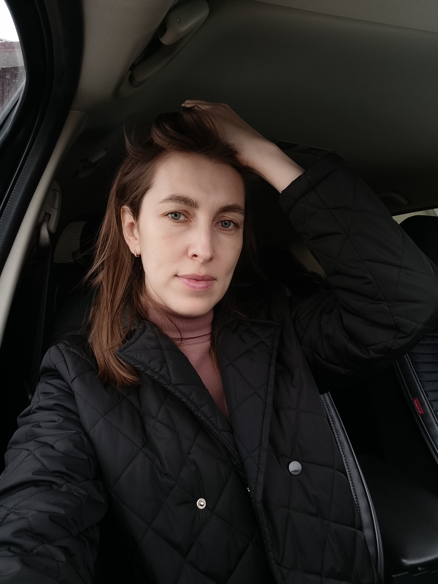 A woman is in the front seat of a car. She is wearing a black quilted jacket over a light pink turtleneck. She is touching her hair with her right hand. The interior of the car is dark, with black seats and trim. The woman has fair skin, light brown hair, and blue eyes. The image appears to be a selfie taken by the woman. There is no evidence or indication of this photo being taken in Фоки, Russia.