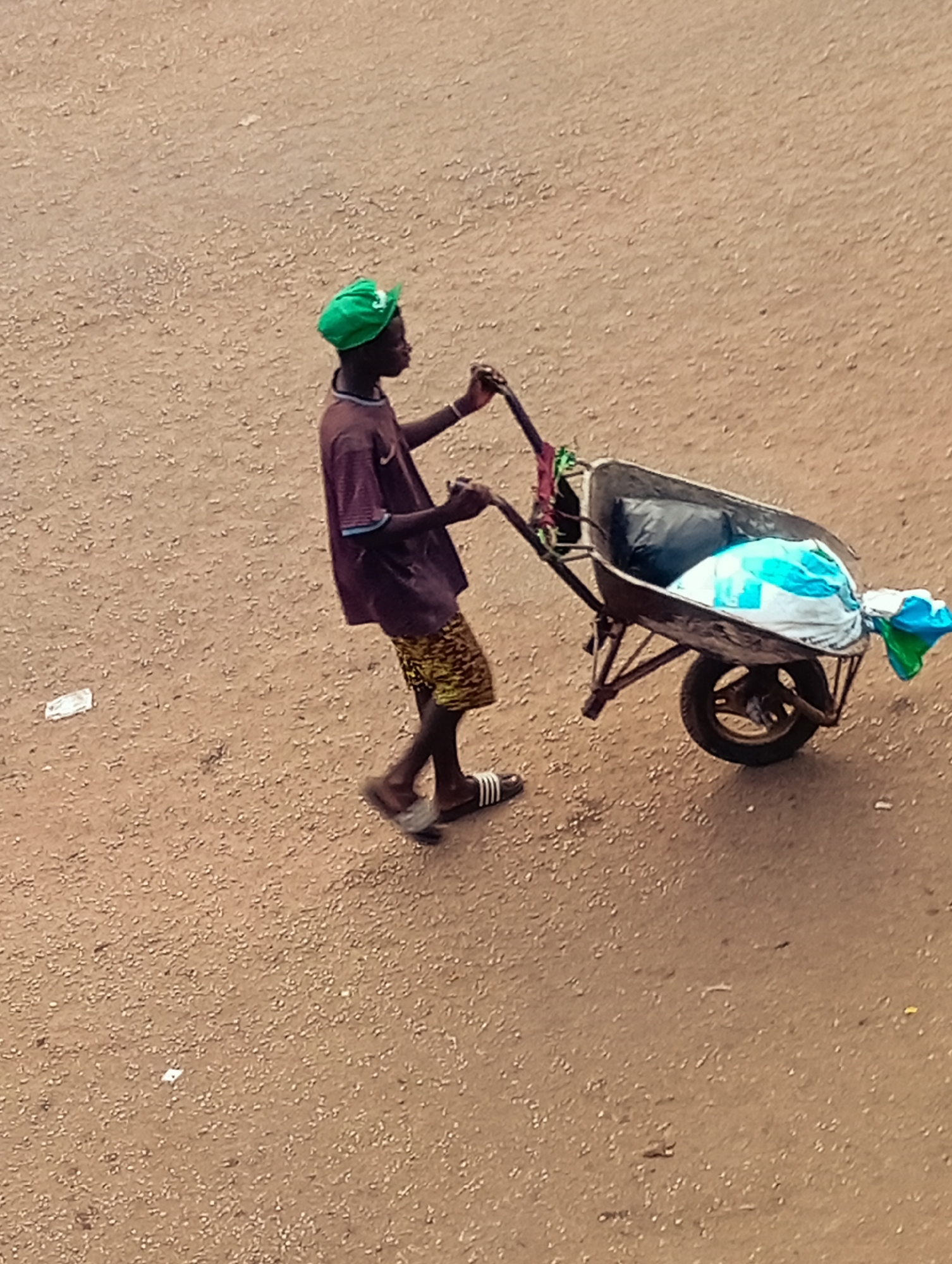 A person wearing a green baseball cap, a brown t-shirt with blue accents, yellow patterned shorts, and black and white sandals is pushing a wheelbarrow. The wheelbarrow is filled with black and blue plastic bags. The scene appears to be Bukuru, Nigeria; the ground is sandy.