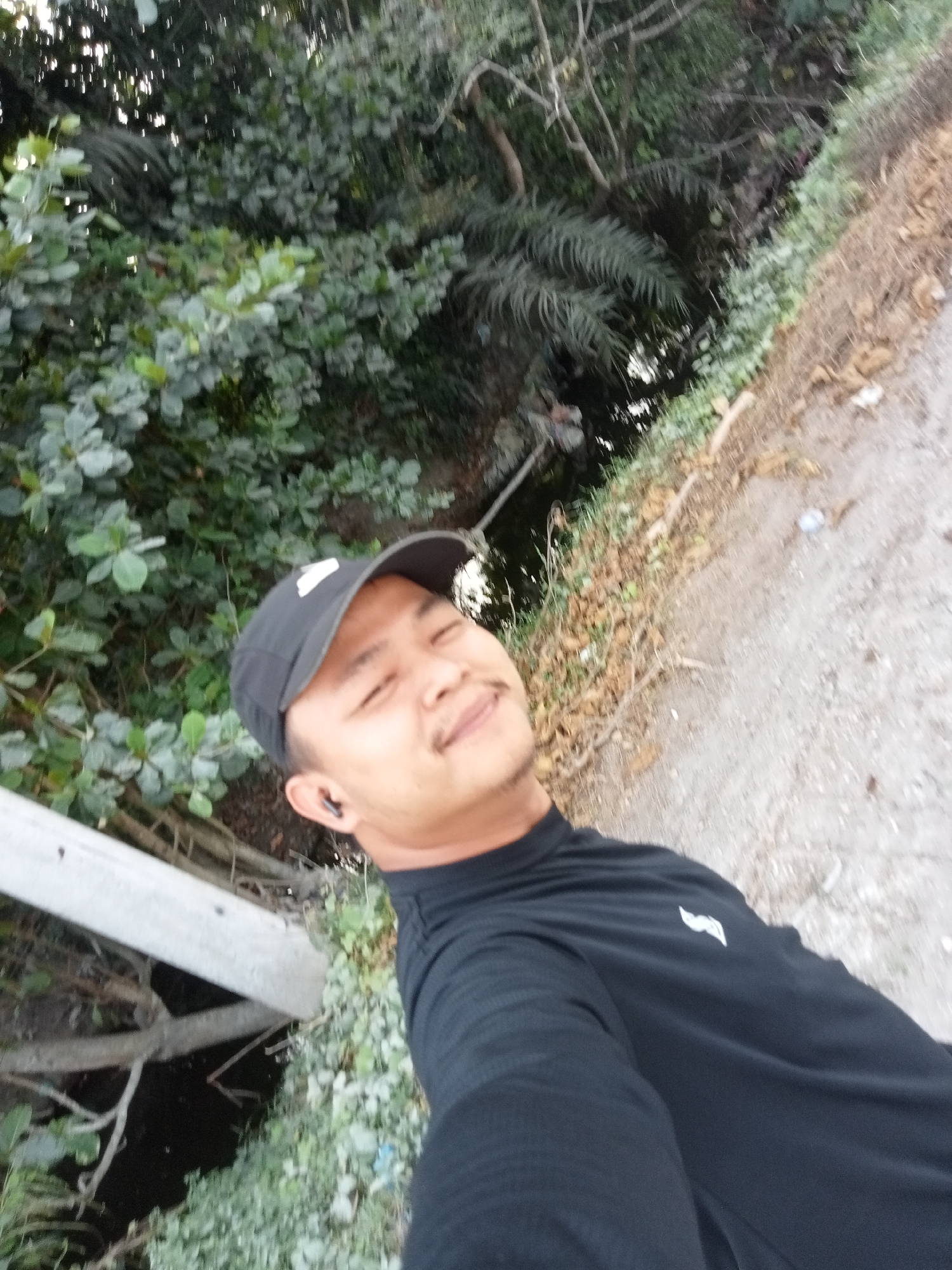 A man wearing a black baseball cap and a black long-sleeved shirt is taking a selfie outdoors. He is positioned on the right side of the frame, with a smiling expression. The background consists of dense green foliage on the left, and a dirt path with dry leaves and some debris on the right. A concrete structure, possibly a bridge railing, is visible in the lower left portion of the image. The lighting suggests it is daytime. The specific location of Teluk Kenidai, Indonesia, is not visually discernible from the image content.