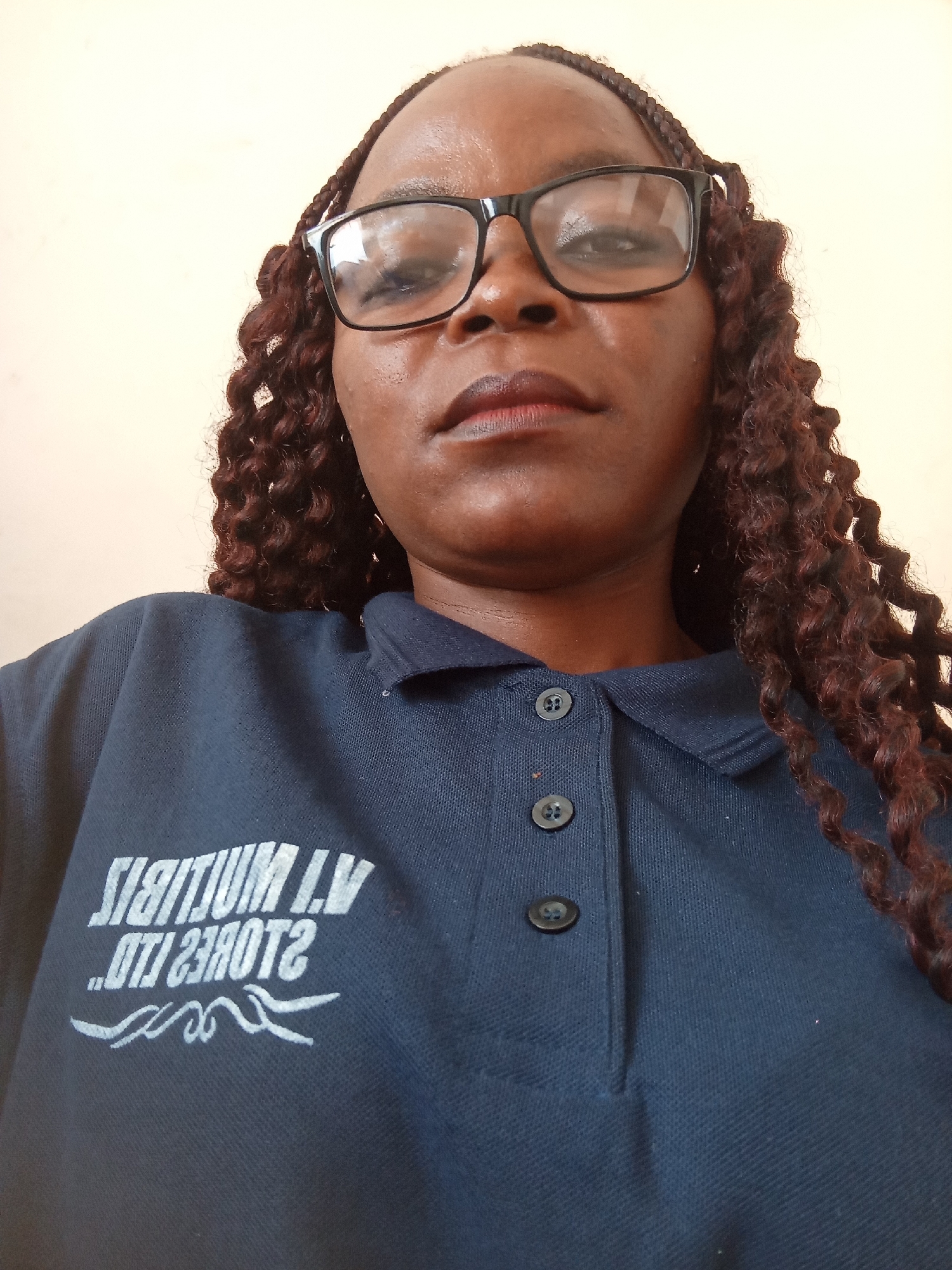 A close-up, waist-up shot shows a dark-skinned woman wearing glasses and a navy blue polo shirt. The woman has braided hair. The shirt has two buttons at the collar. A white logo is visible on the left side of the shirt. The background is a plain, light-colored wall.