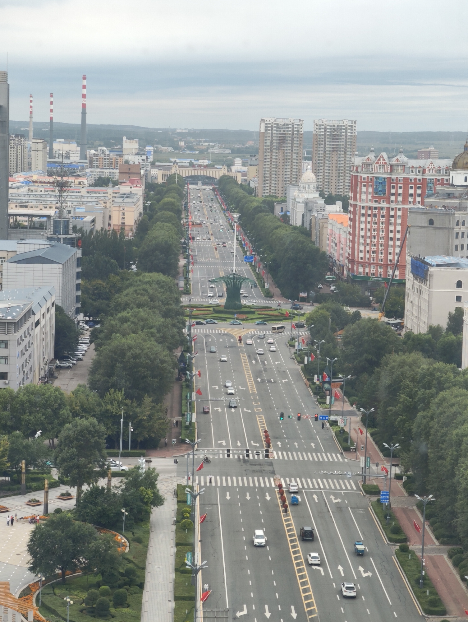 An aerial, eye-level view of a wide, multi-lane city street lined with trees and buildings in 幸福乡, China. The street appears to be a major thoroughfare, with traffic moving in both directions. Buildings of various architectural styles and heights flank the street, including modern high-rises, residential buildings, and some older structures. A prominent feature is a large traffic circle or roundabout in the distance, with a green sculptural element at its center. Pedestrians are visible on sidewalks and crossing the street at designated crosswalks. There are also numerous vehicles on the road, including cars, buses, and possibly trucks. Red flags are visible along the street, decorating lampposts and some median barriers. The sky is overcast, suggesting a cloudy or hazy day. The overall scene depicts a bustling urban environment.
