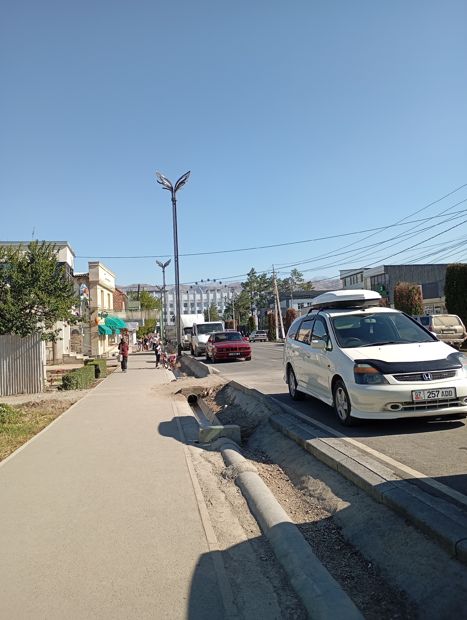 The image depicts a street scene in Talas, Kyrgyzstan. A white minivan with a roof rack is parked on the right side of the road. The minivan has a license plate that reads "07 257 ADD KG". A red sedan is driving in the opposite direction on the left lane. In the background, several buildings are visible, including a multi-story white building with a dark roof, and other smaller commercial buildings. A lamppost with two lights stands on the sidewalk to the left of the road. The sidewalk is paved and runs alongside the road. There are several people walking on the sidewalk. A drainage ditch runs along the edge of the sidewalk, with pipes and construction materials visible. The sky is clear and blue, indicating a sunny day. The overall scene suggests a typical urban environment with traffic, pedestrians, and infrastructure.