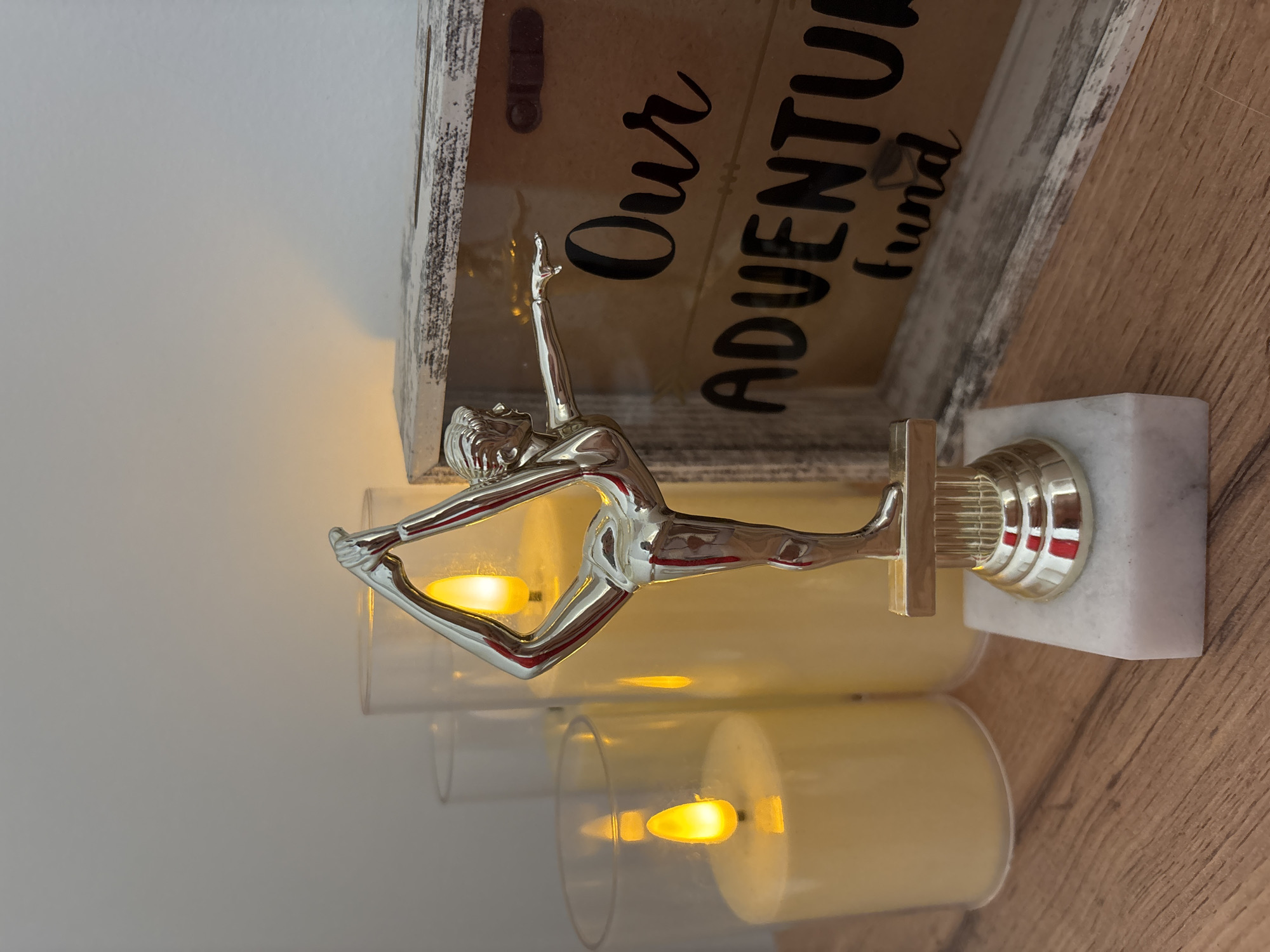 An artistic, silver-toned figurine of a woman in a yoga pose stands on a marble base. Behind the figurine, three tall, unlit pillar candles are encased in clear glass cylinders. To the right of the figurine is a rustic wooden box with a glass front. The box has the words "Our Adventure Fund" painted on it in black cursive letters. The items are arranged on a wooden surface. The image is well-lit, with a warm glow emanating from the candles. There is no indication of the location being in Osypnoi Bugor, Russia.