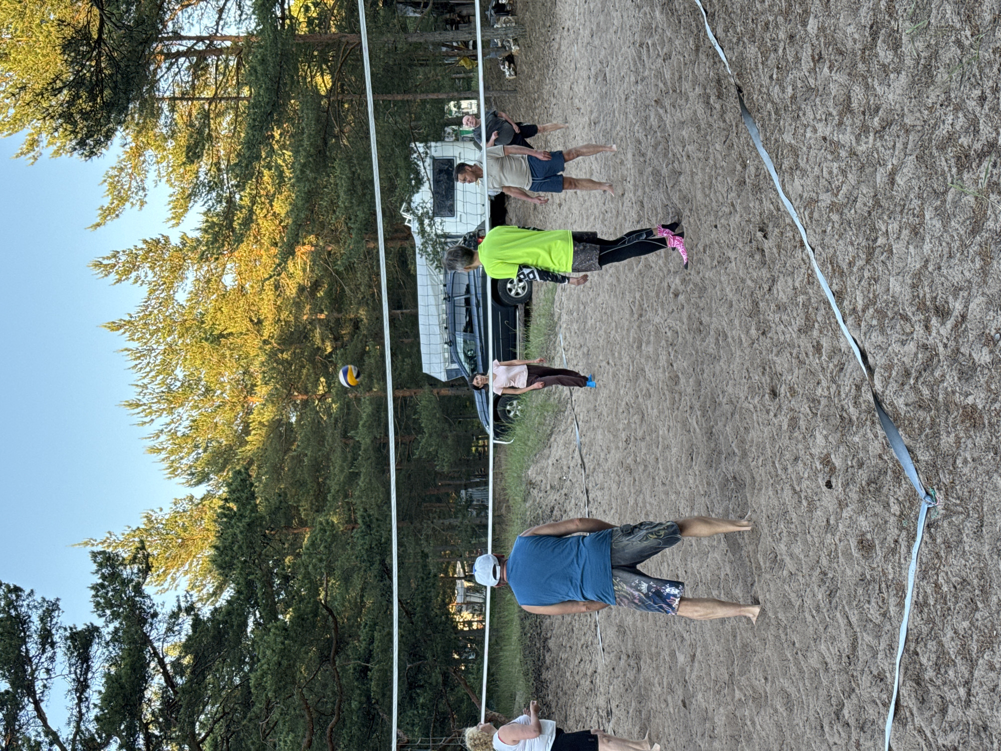 In Zelenaya Roshcha, Russia, a group of six individuals is engaged in a game of beach volleyball on a sandy court. The court is marked with white tape, and a volleyball net stretches across the playing area. The players, comprising three men and three women, are dressed in casual athletic wear. A volleyball is in mid-air, suggesting an active moment in the game. The background features a dense forest of pine trees, a camper van, and a dark-colored car, indicating a natural, possibly recreational setting within or near Zelenaya Roshcha. The sky above is clear and blue, and the lighting suggests it is either late afternoon or early evening.