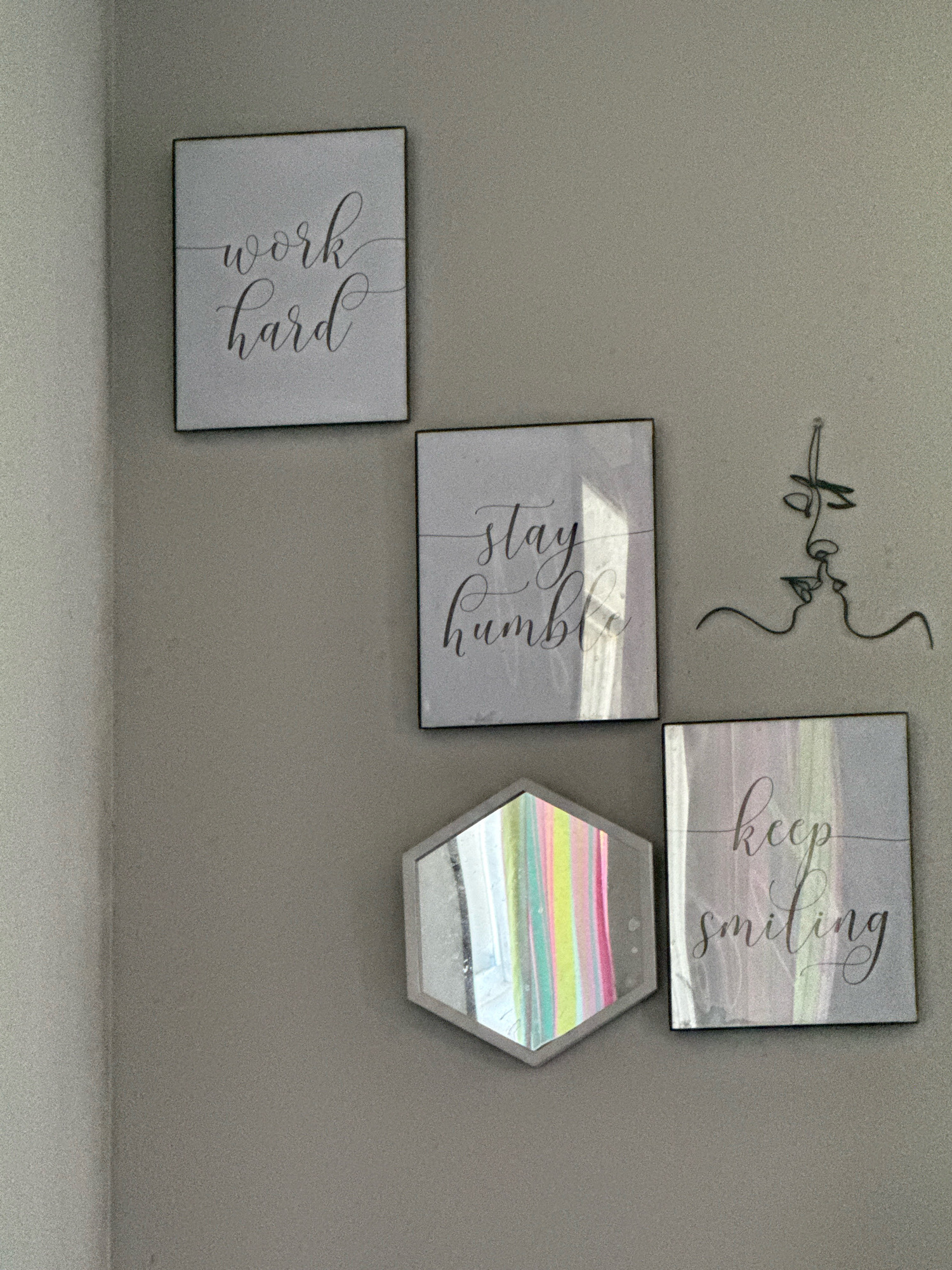 The image shows a wall with several decorative items. There are three framed pieces of art with cursive text, reading "work hard", "stay humble", and "keep smiling". To the right of the "stay humble" artwork is a metal wire sculpture of two faces kissing. Below the "stay humble" artwork is a hexagonal mirror with colorful stripes reflected in it. The wall is a light gray color, and there is a slight shadow cast by a corner on the left side of the image. The scene appears to be an interior decoration in a home. There is no indication of the location being in Mora, United States of America.