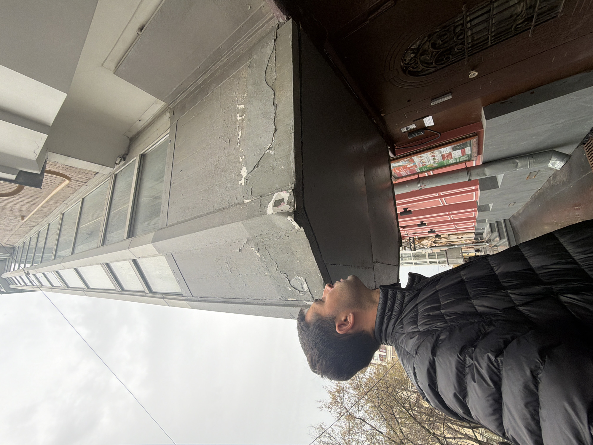 A man in a black puffy jacket looks up at the corner of a building in Moscow, Russia. The building is gray with white trim and multiple windows. Above the man is a dark wooden door with a decorative iron grill. Nearby are trees with bare branches, under a cloudy sky. In the background, signs of the city, including other buildings and signs, are visible.