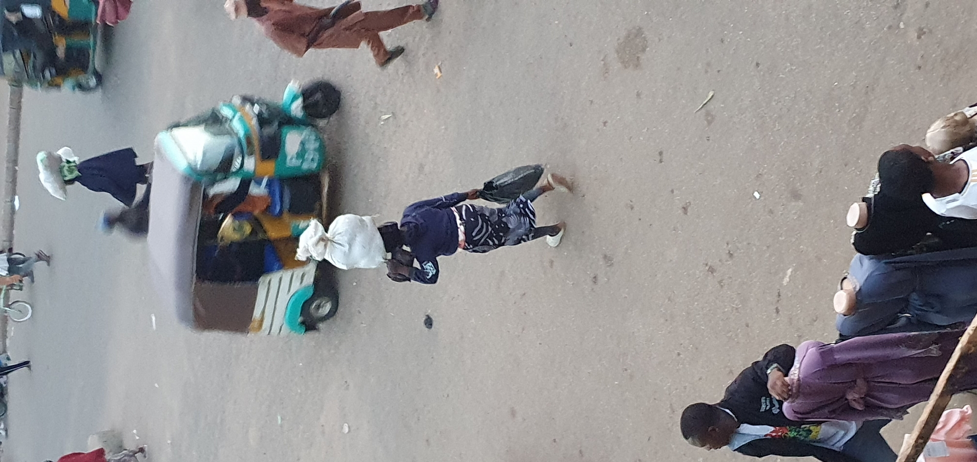 The image depicts an outdoor scene, likely a street or market, with several people and vehicles. The perspective is from above, looking down at an angle. The ground is paved and appears to be a street with some scattered debris.
In the foreground, a person wearing a dark blue long-sleeved shirt and patterned pants is walking. They are carrying a dark bag in their right hand and have a white bag balanced on their head. To their left, a colorful three-wheeled vehicle, possibly a tuk-tuk, is parked. This vehicle is loaded with various items, including what appear to be fabrics or bags.
Further in the background, another person in a brown outfit is walking away from the viewer. To the right of the image, a group of people are gathered, with some seated and others standing. One person in the foreground on the right is wearing a white shirt with dark trim. Another person next to them is wearing a dark garment and a purple cloth draped over their lap.
The lighting suggests it is daytime, likely late afternoon or early morning, given the angle of the shadows. The weather appears to be clear, with no visible rain or fog. The overall atmosphere seems busy, characteristic of a public thoroughfare or marketplace where people are transporting goods or moving about their daily routines. There is no visible text in the image.