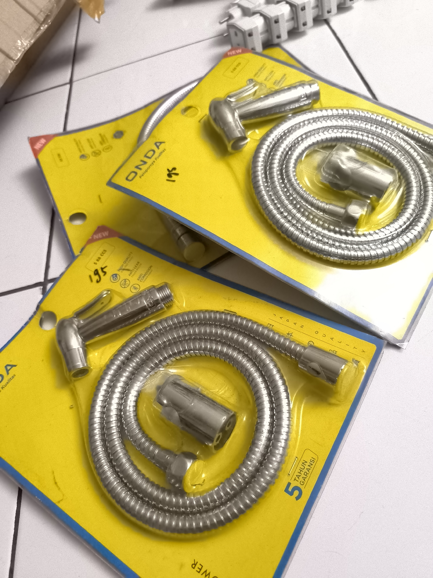 Two packages of ONDA bidet sprayers are displayed on a tiled surface. The packaging is bright yellow with blue borders. The top package, slightly to the left and angled, shows the brand name "ONDA" and some text in Indonesian. A price of "195" is visible on this package. The bidet sprayer and hose are packaged in clear plastic. The bottom package is similar, also displaying the ONDA branding and an Indonesian slogan "Pelopornya Kualitas". This package also shows "5 TAHUN GARANSI" which translates to "5 YEAR WARRANTY." A white electrical strip with multiple outlets is visible in the background, out of focus. The surface appears to be a light-colored tiled floor.