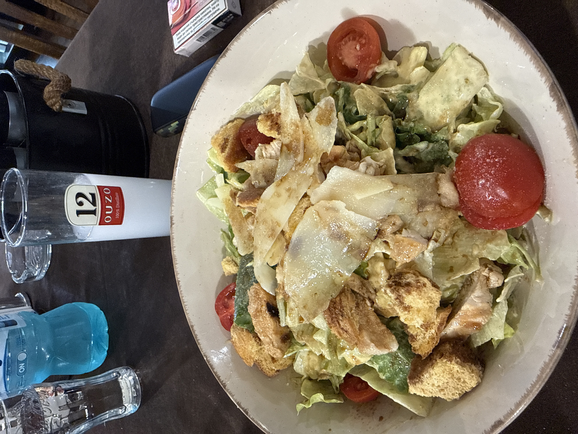 The image shows a Caesar salad in a white ceramic bowl. The salad includes lettuce, croutons, tomatoes, and parmesan cheese. The salad appears to be dressed with a creamy dressing. Next to the bowl is a bottle of water and a bottle of Ouzo 12 with a glass. A pack of cigarettes can be seen. The scene appears to be set on a table in a restaurant. No information about the city of Lukovit, Bulgaria can be found in the image.