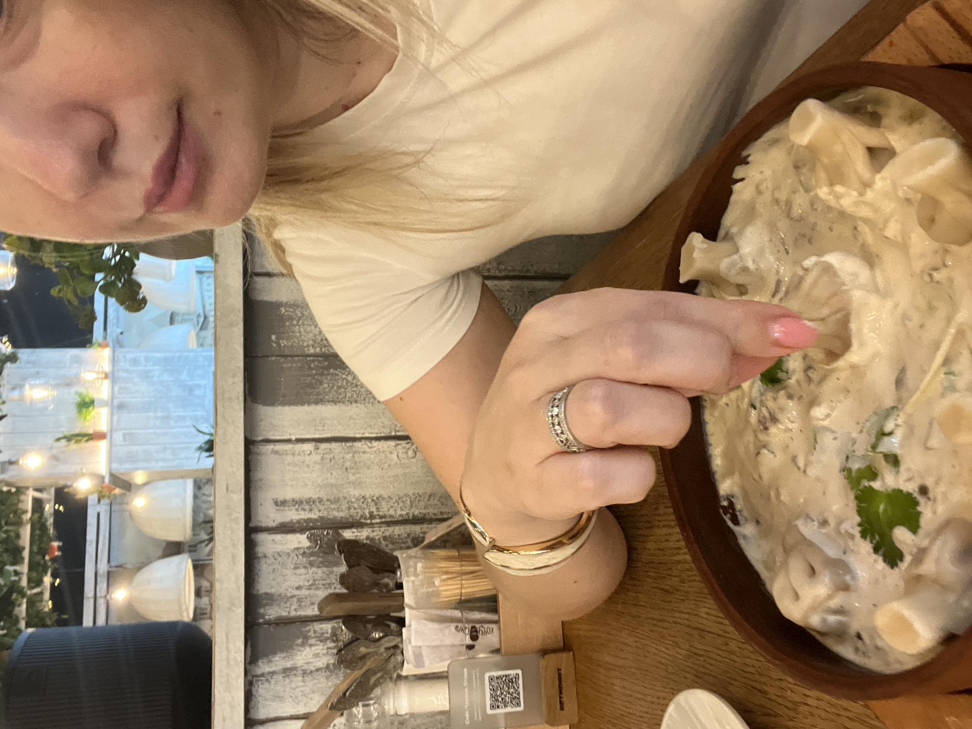 A woman with blonde hair and pink nail polish is sitting at a table and reaching for food in a brown bowl. The food appears to be dumplings in a creamy sauce with a garnish of cilantro. She is wearing a white t-shirt and several bracelets on her left wrist, along with a ring on her left ring finger. In the background, there is a rustic wooden wall, a black cylindrical object, and decorative pendant lights. The scene is set in a restaurant in Sochi, Russia.