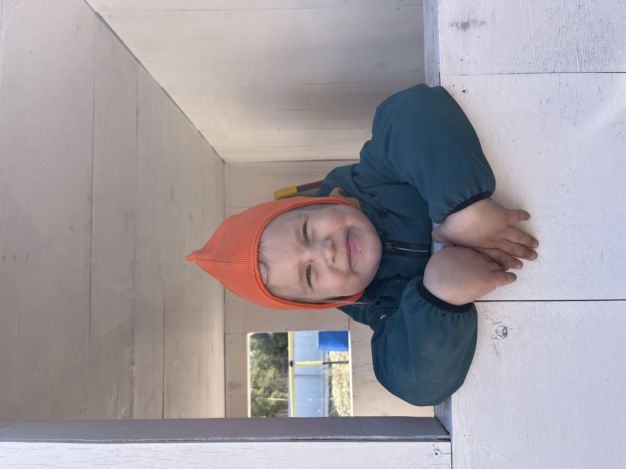 A child wearing an orange knit cap and a dark teal jacket is shown indoors in a bright, white-painted wooden structure with an opening that looks outside to a playground. The child is smiling and leaning forward with their hands resting on the edge of the structure. The playground, visible through the opening, shows trees, a yellow metal structure, and a light-colored fence. The location is Chaikovsky, Russia.