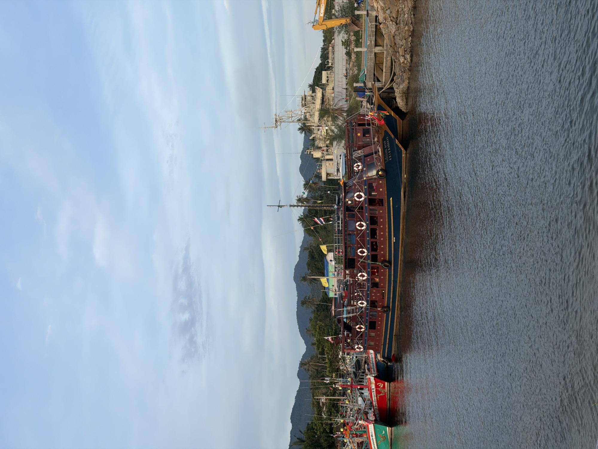 The image depicts a waterfront scene in Koh Phangan, Thailand, likely a harbor or a pier. Several boats are moored in the calm, dark water, with a rocky embankment visible on the left. The largest boat, with a blue and maroon hull and gold trim, is prominently featured, displaying life preservers along its side. It appears to be a passenger or tour boat, with multiple windows and decks. Smaller fishing boats, some painted in vibrant red and green, are docked alongside.
In the background, the shoreline is dotted with lush green palm trees and vegetation, leading up to misty, blue mountains. On the land side, construction equipment, including a yellow excavator, suggests ongoing development or maintenance work. A few people are visible near the construction area and on the boats, engaged in what appears to be routine activities rather than a specific event.
The sky is a soft, hazy blue with scattered white clouds, indicating it might be late afternoon or early morning. The overall atmosphere is peaceful and serene, with the gentle ripples on the water contributing to the tranquil mood.
Visible text includes Thai script on the side of the largest boat, near its stern, though it is difficult to fully decipher from the image. The Thai text on the side of the large boat reads "บุญมาศ 40422".